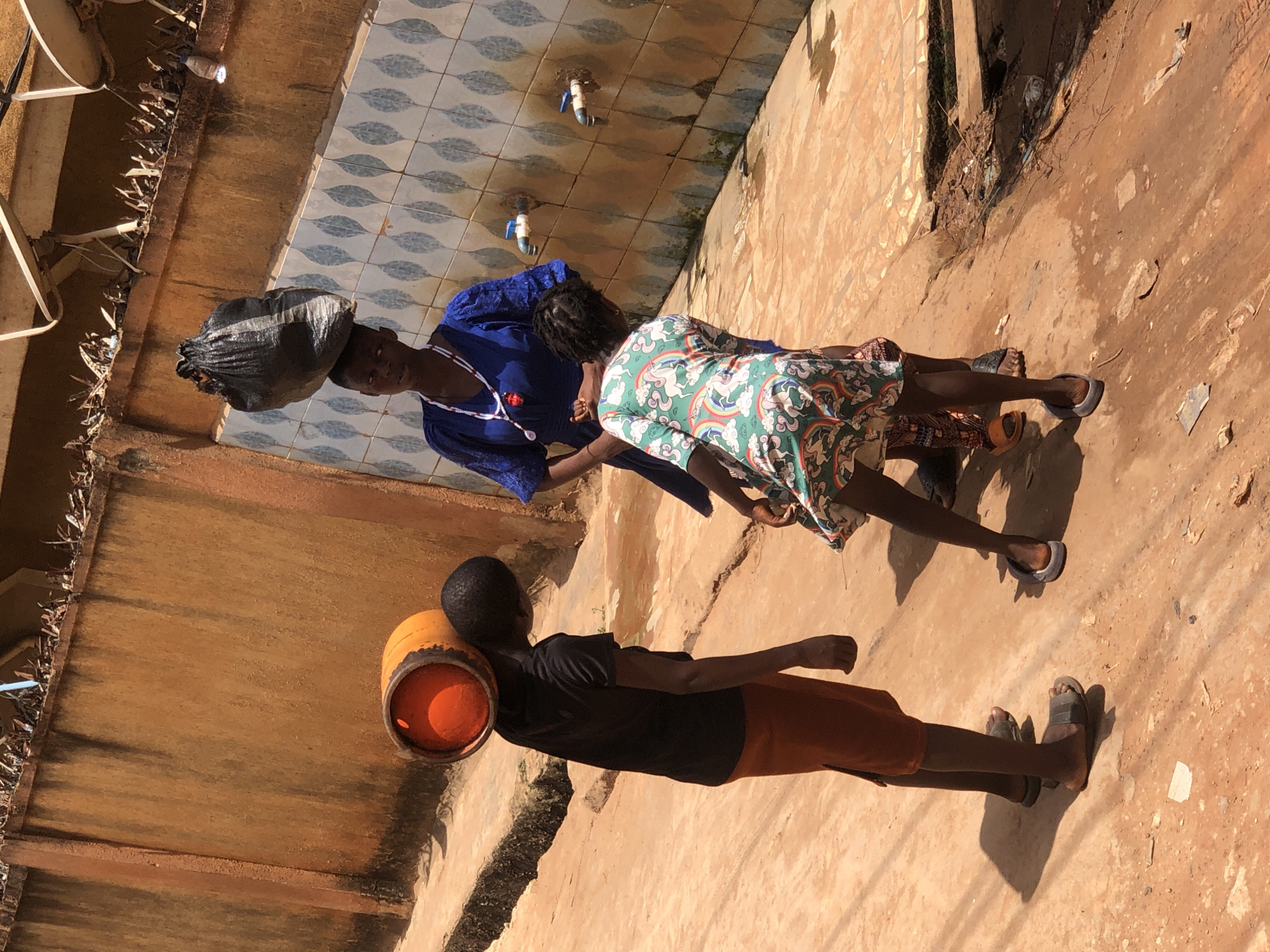 The image shows three people in an outdoor setting in Onitsha, Nigeria. A woman wearing a blue dress and a white necklace carries a large black bag on her head. Standing next to her is a child in a green and white dress. A boy wearing a black shirt and orange shorts carries an orange container on his shoulder. They are standing on a dirt ground next to a building with tiled walls and water spouts. The location appears to be a residential area.