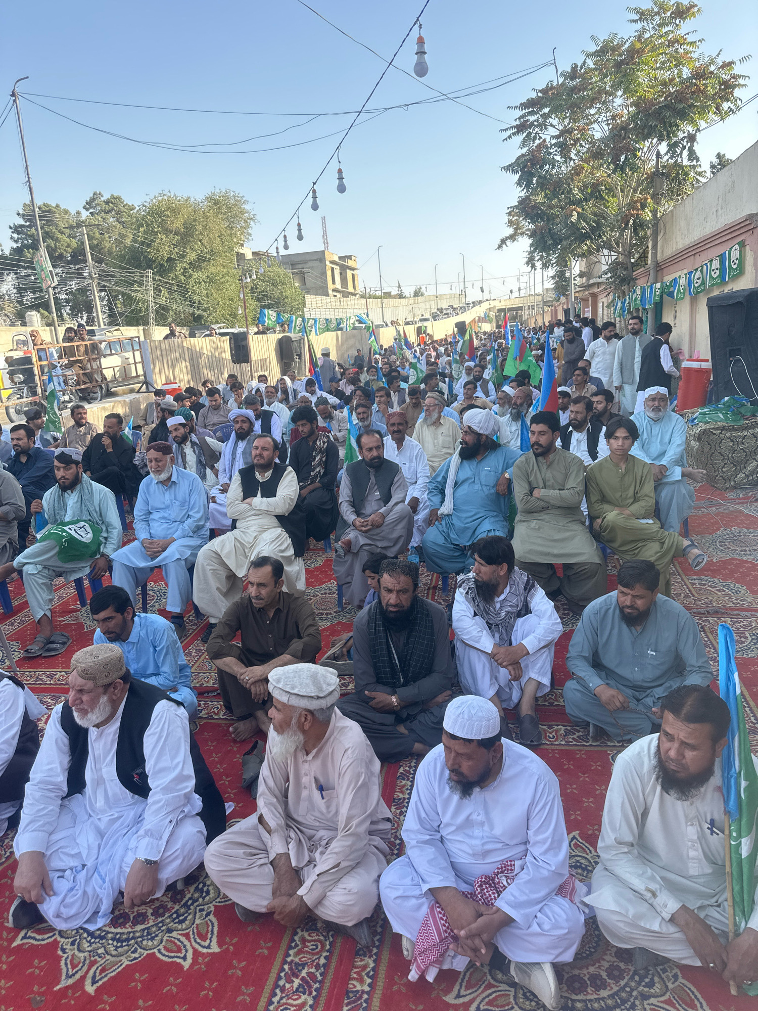 An outdoor gathering is taking place on a red carpet, likely in the city of Quetta, Tehsil, Pakistan. A large crowd of men, dressed in traditional South Asian attire, are seated on the ground, some on blue chairs. Many are wearing white or light-colored shalwar kameez and some have turbans or caps. Several men are holding flags, predominantly green and blue, with some featuring a crescent moon and star symbol.
In the foreground, several men are prominently featured. One man on the far left is wearing a white shalwar kameez with a black vest and a white cap. Next to him, another man in a similar white outfit is looking to his right. To his right, a man in a light-colored shalwar kameez is seated with his hands in his lap. Further to the right, a man in a light green outfit is sitting with his arms crossed. Behind them, a man in a white shalwar kameez and a light-colored vest is seated, and another man in a light brown outfit is also seated.
In the background, the gathering extends further back, with more people visible. Banners with text and symbols are displayed along a wall or fence. Trees and buildings are visible beyond the crowd, and a clear sky suggests it is daytime. Electrical wires and poles are also present, indicating an urban or semi-urban setting. The overall atmosphere appears to be that of a political rally or a community event.