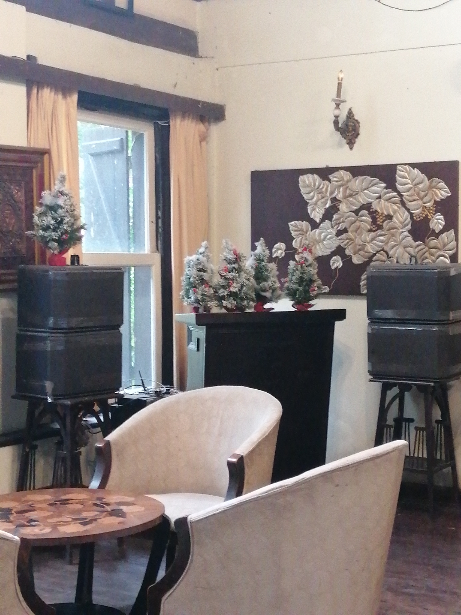 The image captures a cozy and festively decorated indoor space, likely a cafe or lounge, in Da Lat, Vietnam. No people are visible, contributing to a tranquil atmosphere.
In the foreground and mid-ground, two light-colored upholstered armchairs with dark wooden frames are arranged around a small, round wooden table. The table features an ornate wooden inlay pattern, adding a decorative touch. On either side of the room, dark wooden stands support stacks of two dark, rectangular boxes, which appear to be speakers, hinting at an entertainment setup.
The background features a large window, framed by light-colored curtains, allowing natural daylight into the room and offering a view of greenery outside. Above the window, dark wooden beams accent the ceiling. A dark console table or mantelpiece stands against the back wall, below a large dark artwork adorned with embossed silver-colored leaf and berry patterns. A decorative wall sconce with an artificial candle is mounted above the artwork.
Several small, frosted artificial Christmas trees, decorated with red berries and snow-like accents, are placed on the console table and a stand near the window, suggesting a holiday or festive season. The overall soft lighting indicates a peaceful daytime setting. The decor blends elements of traditional elegance with a festive, comfortable ambiance.