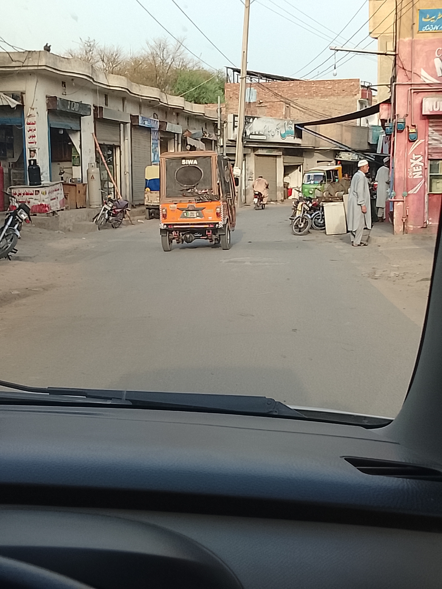 The image shows a street scene in Nasheman Colony, Pakistan. The view is from inside a car, looking out through the windshield. In the street, an orange SIWA auto-rickshaw is moving away from the camera. Several motorcycles are parked on the sides of the road. A man wearing traditional Pakistani clothing is standing near a shop on the right side of the street. The buildings lining the street are a mix of shops and residences. Power lines are visible overhead. The weather appears overcast.