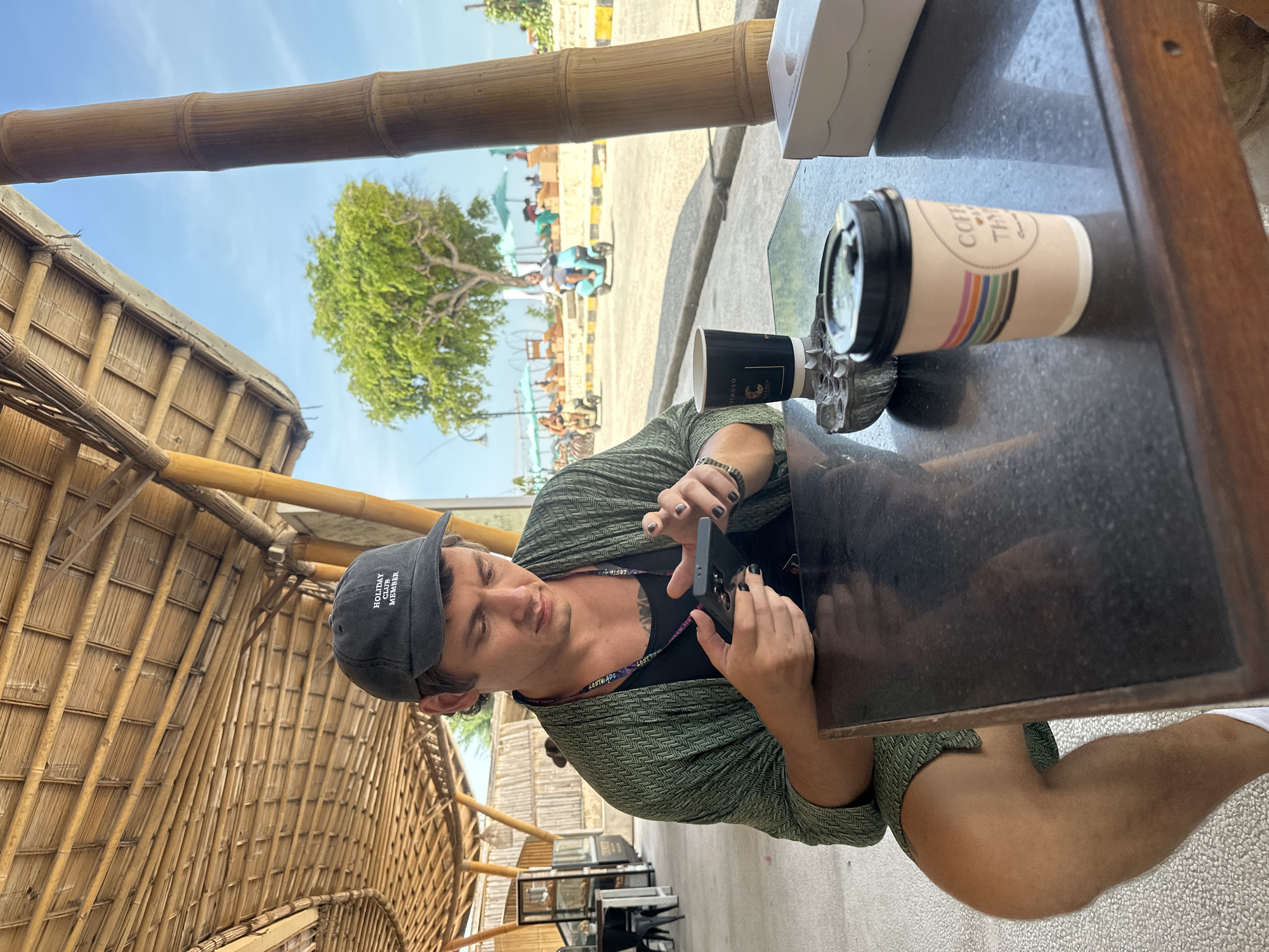 A horizontal medium shot depicts a man sitting at a table in an outdoor cafe. He is wearing a black t-shirt, a green patterned shirt, a grey baseball cap with "Holiday Club Member" text, and a lanyard with "LOST TAPE" on it. He is holding a smartphone in his hands and looking at it with a focused expression. His nails are painted black. A coffee cup with a colorful stripe design sits on the table to his right, next to a black coffee cup. A white box is also on the table. The table is made of dark marble.
The cafe appears to be constructed primarily of bamboo, with bamboo framing visible above and to the left of the man. In the background, beyond the cafe, there is a street scene with people, vehicles, and a tree. The location is Unknown, Indonesia.