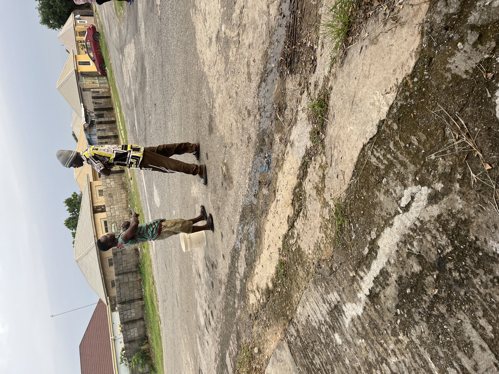 In an outdoor, urban setting in Abuja, Nigeria, two young men stand on a paved road. The man on the left wears a green and red striped sleeveless shirt, tan shorts, and dark sandals, with a white bucket to his left. He appears to be gesturing with his right hand. The man on the right is dressed in a patterned short-sleeved shirt with yellow, black, and white designs, brown pants with white stripes, and a grey beanie. He has his arms crossed. The road is weathered and shows patches of discoloration and puddles. In the background, residential buildings with light-colored walls and silver or brown roofs are visible, separated by low concrete block walls. A red car is parked on the side of the road. The sky is overcast.