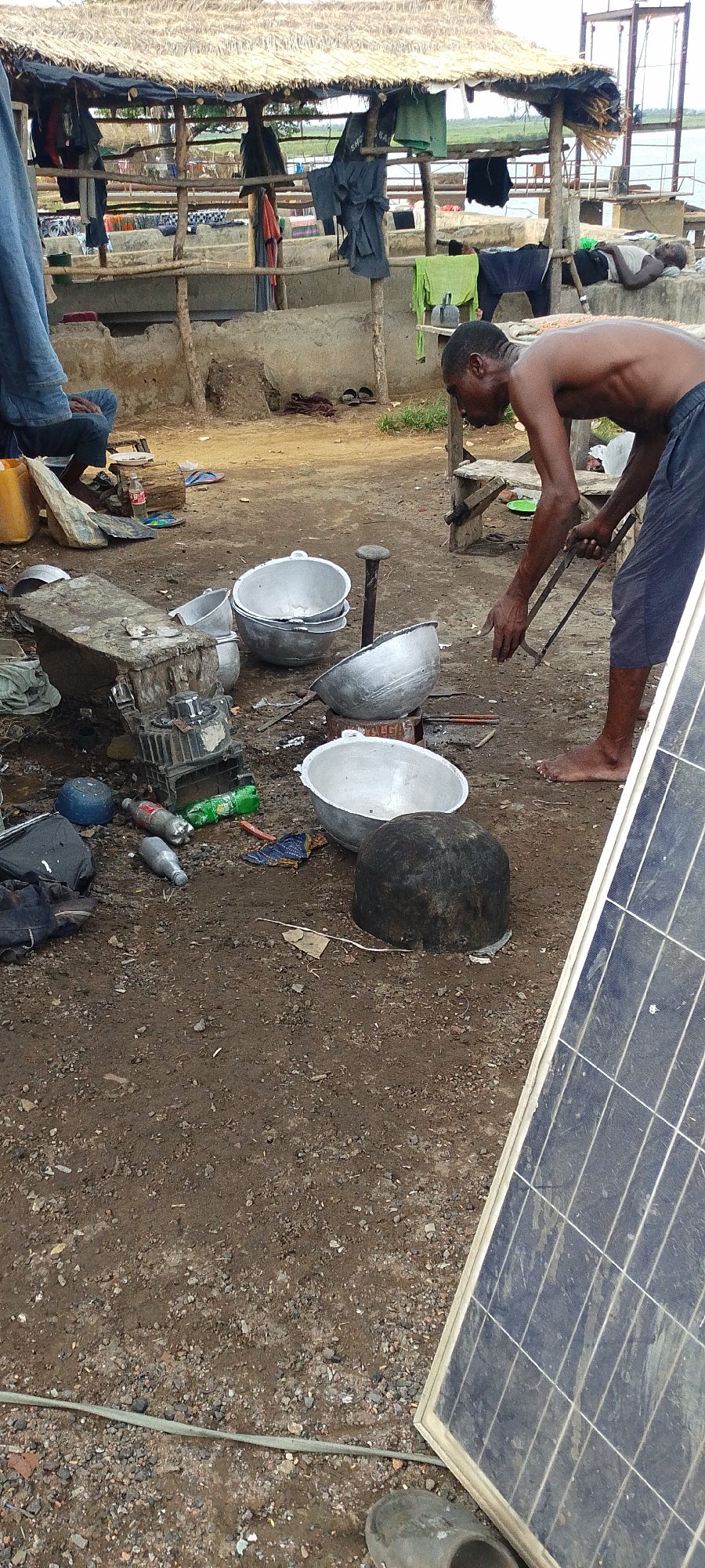 In Lokoja, Nigeria, a shirtless man wearing navy shorts uses metal tongs to handle a cooking pot placed over a burning stove. Several aluminum pots surround him. Various items, including plastic bottles, engine parts, and scraps, litter the ground around his feet. A solar panel leans against the ground nearby, and part of a thatched roof structure is visible in the background, with people and clothes present. Concrete structures are also in the background, close to a river.