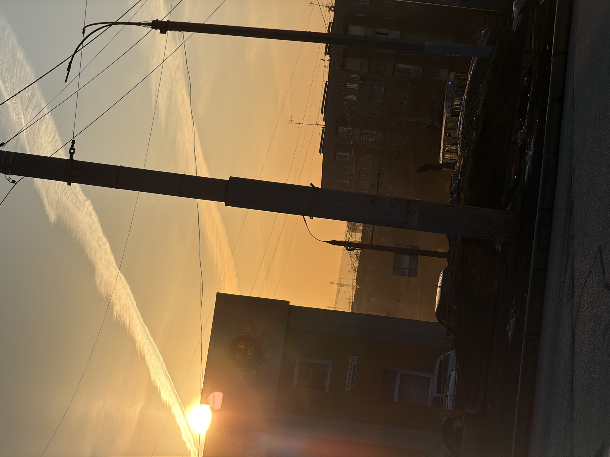 The image captures a cityscape at sunset in Izhevsk, Russia. The sun is low on the horizon, casting a warm golden light across the scene. A prominent contrail stretches across the sky from left to right. Several utility poles and wires crisscross the foreground. A building on the left displays a digital clock with the time 18:30 and a temperature of 16°C. The street sign on the building reads "Yakurtsky 16". A multi-story apartment building is visible in the background. A dark, uneven surface, likely a sidewalk or street, runs along the right side of the frame. Cars are parked on the right.
