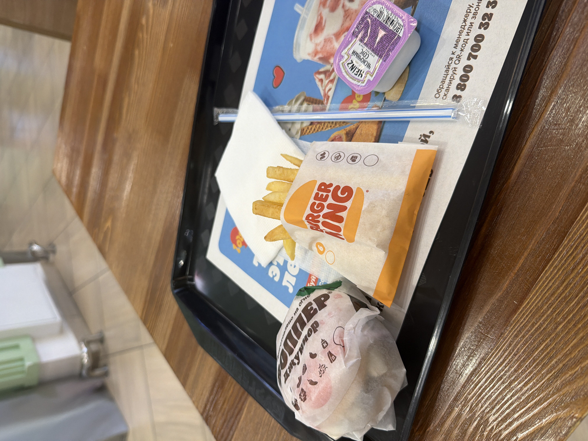 The image shows a Burger King meal on a black tray on a wooden table. The meal consists of a burger wrapped in white paper with the Burger King logo, a bag of french fries, and a small container of garlic sauce from Heinz. The burger wrapper has illustrations of food items and a pink face. A straw is visible extending from under the french fry bag. The background includes part of a blue tray liner with a cartoon image of a cow and some text. The text on the tray liner includes a phone number and instructions to contact a manager or scan a QR code. The overall scene appears to be a casual meal setting, likely indoors. The lighting suggests it could be daytime, but no specific time can be determined. The image is taken from a slightly overhead perspective, focusing on the food items.