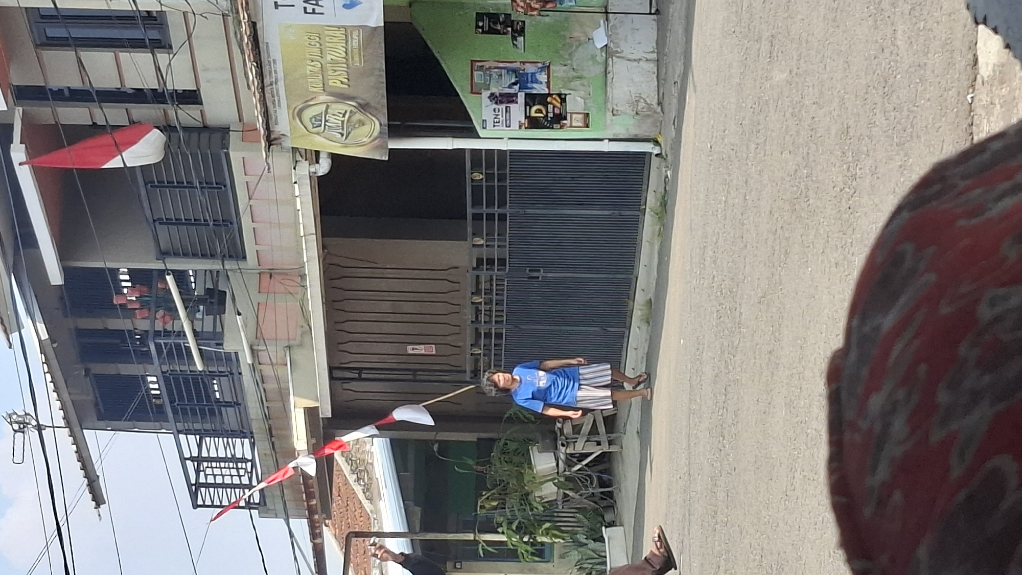 A medium shot captures a woman walking on a paved street in Cilaku, Indonesia. She is wearing a blue t-shirt and striped shorts, with flip-flops on her feet. The woman is the primary subject, walking towards the right side of the frame. Behind her, a dark metal fence encloses a property. To the left, a small plant is visible. In the background, a building displays Indonesian flags, suggesting a celebration or patriotic event. The sky is clear and blue. The overall scene depicts a common street view in the area.