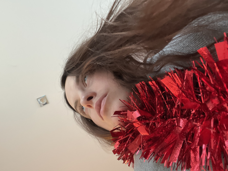 A low-angle, upward-looking photograph depicts an adult female subject indoors. The subject has long, dark brown hair and light-colored eyes, gazing directly upwards towards a plain white ceiling. A dense, shimmering red tinsel garland is draped across the subject's upper chest and neck, partially obscuring the lower face. The subject wears a grey garment, visible beneath the tinsel. In the upper-center of the white ceiling, a single, recessed light fixture, appearing square or circular, is visible. The specific location in the city of Callbach, Germany, or any other geographic detail beyond being indoors, cannot be determined from the visual information provided.