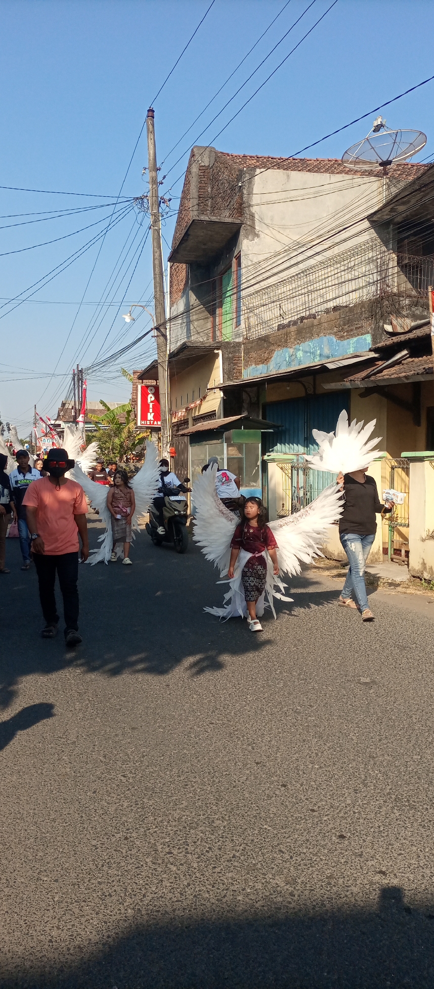 A parade is taking place on a street in Bulungcangkring, Indonesia. Several people are walking in the parade, including children dressed in angel costumes with large white wings. One child is riding on the back of a motorcycle. Buildings line the street, and there are power lines overhead. The sky is clear and blue.