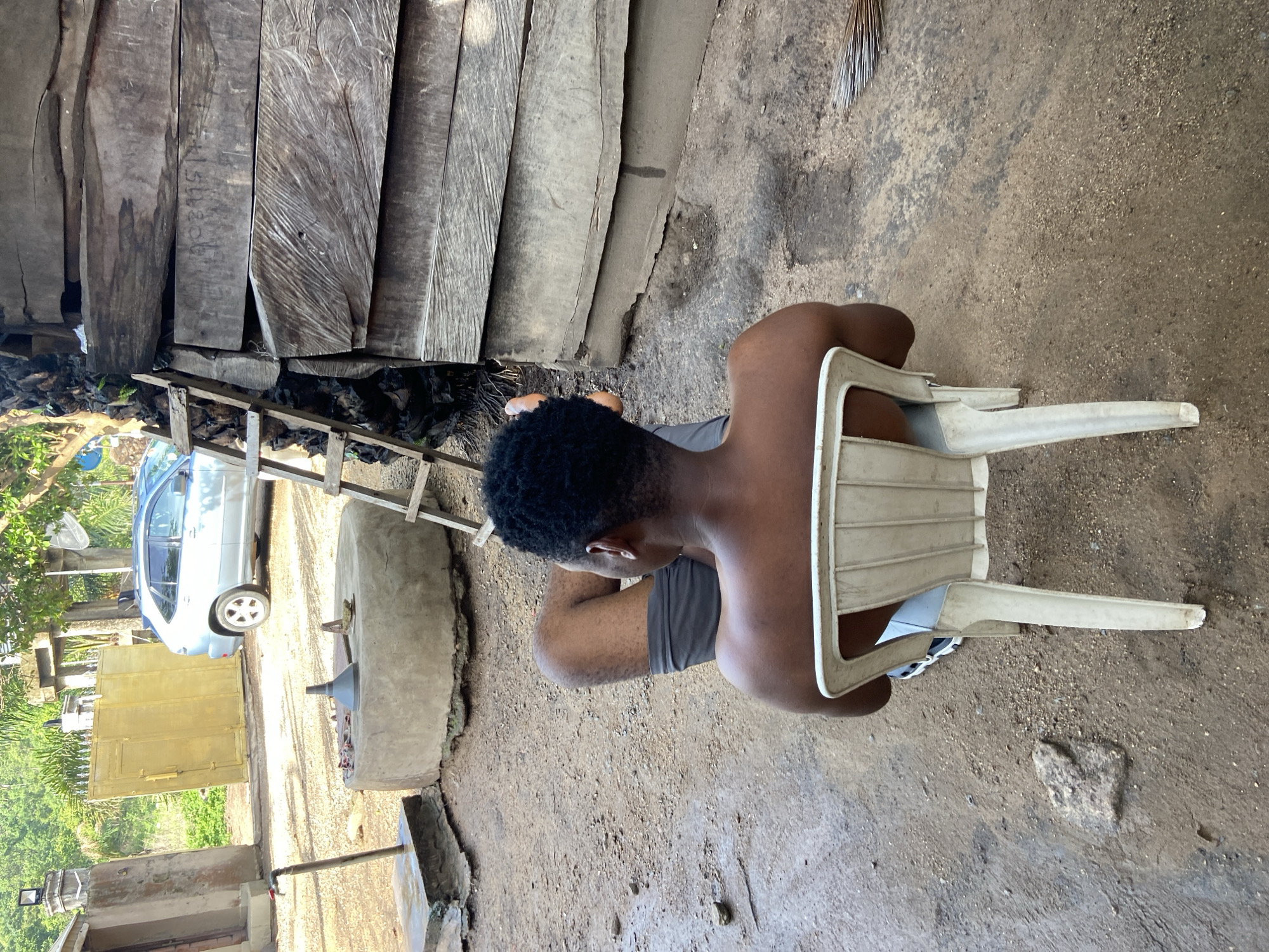 A person with dark hair and skin sits on a white plastic chair in an outdoor area. They wear a grey top and shorts. The ground is dirt and gravel. To the left is a wooden structure, possibly a building, and some vegetation. A silver car is visible in the background, along with a yellow cabinet and what appears to be a traditional milling stone. The location is likely Mararaba, Nigeria, judging by the surrounding environment.