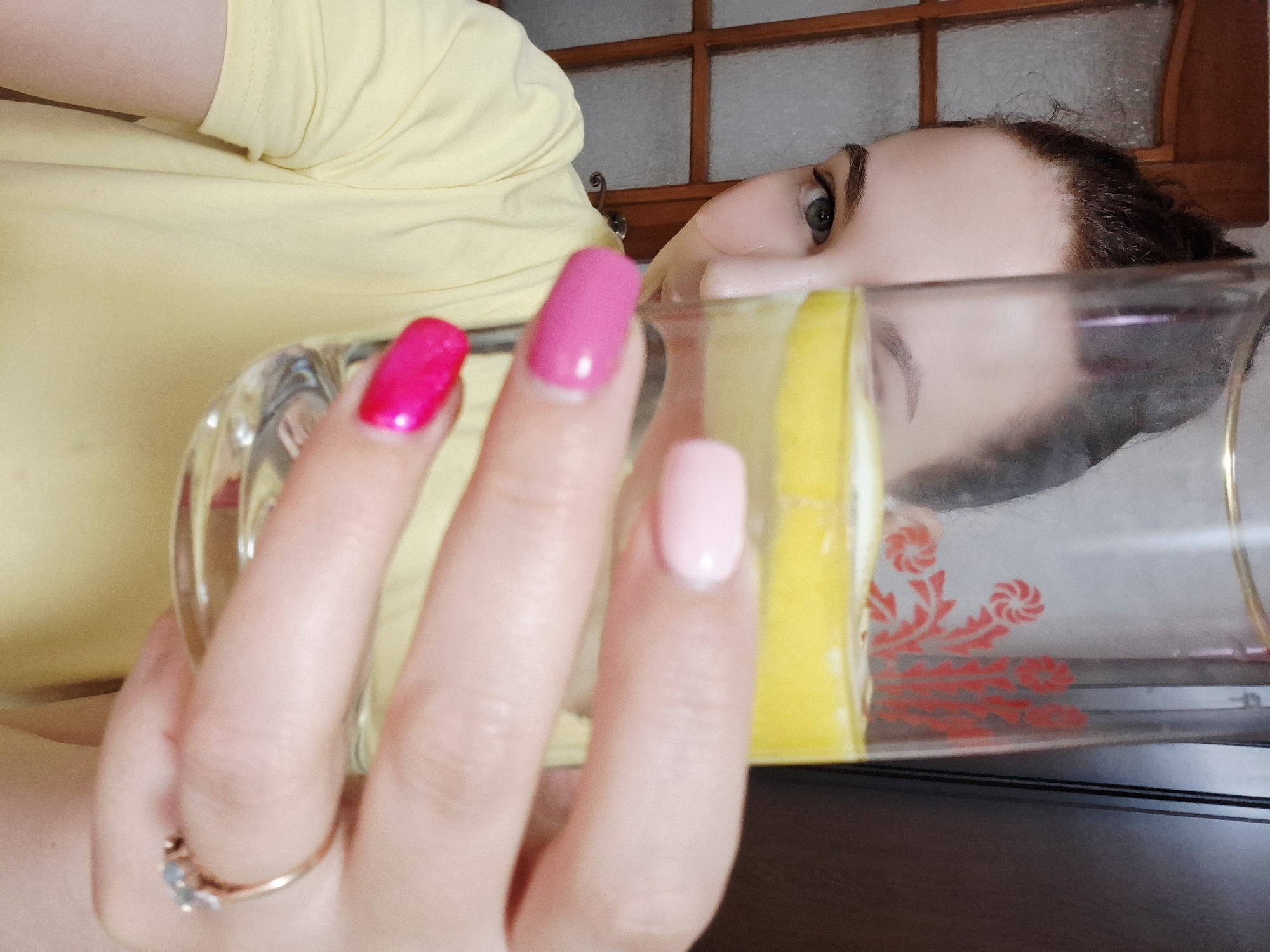 The image shows a person holding a glass with a yellow liquid, possibly lemonade, in front of their face. The person has painted nails of different shades of pink. The reflection in the glass distorts the person's facial features. A ring is visible on the person's finger. The background includes a wooden frame and a dark surface. The presence of the glass and drink is the only indicator of location, without additional details about the city of Chaikovsky, Russia. The overall scene appears to be indoors.