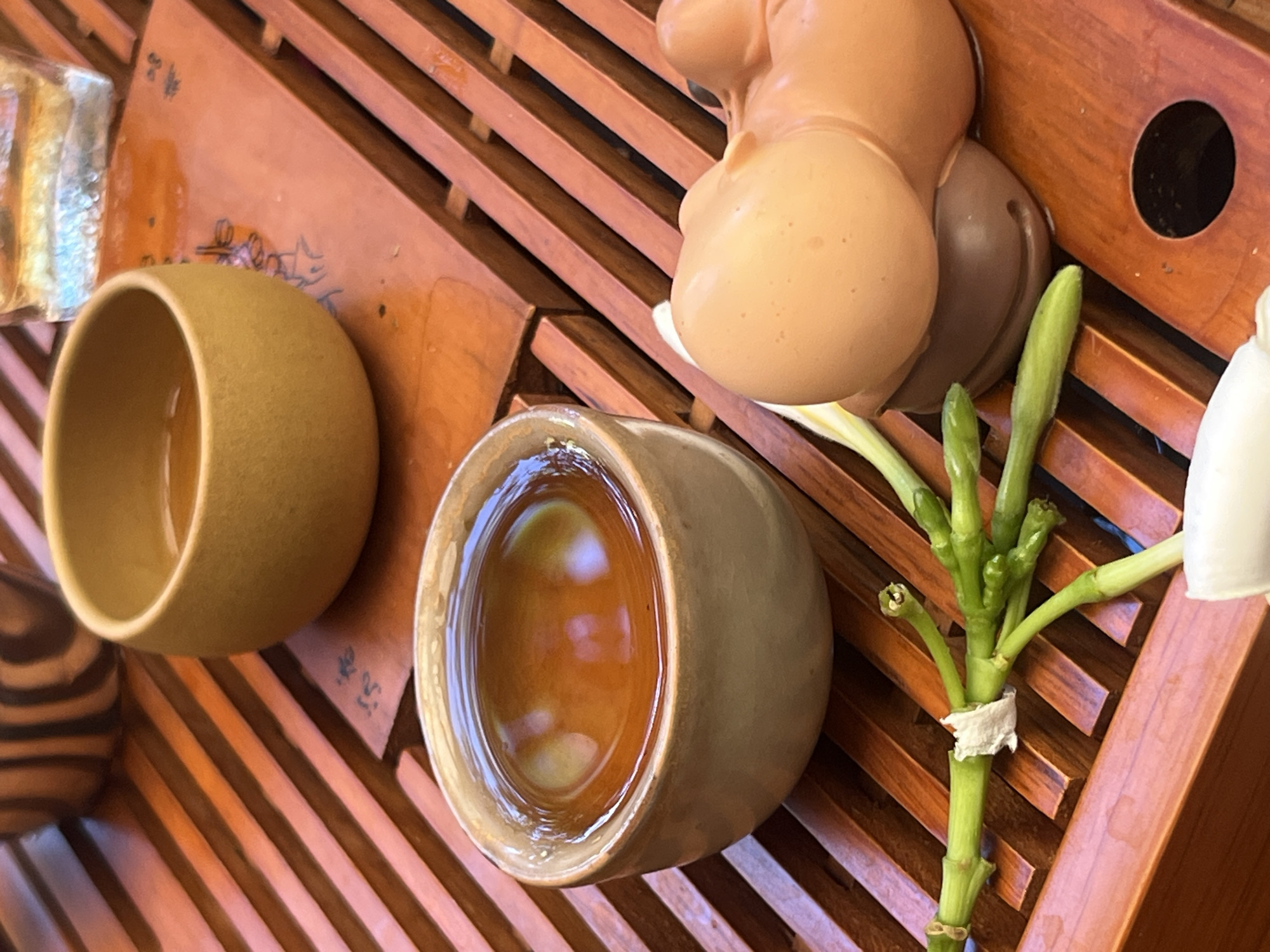 This is a close-up, high-angle shot of a tea set on a wooden tray. The tray has an open-slat design. In the foreground, there is a small cup filled with amber-colored liquid, likely tea. Next to the cup are a few green stems with small buds, tied together with a small piece of white paper. To the right of the stems is a light brown, anthropomorphic sculpture or figurine, possibly made of resin or plastic, resting on a darker brown base. Further back, another small cup, this one a muted yellow, also contains amber liquid. Partially visible in the upper right corner is a clear glass container with ice and liquid, suggesting a beverage. The setting appears to be indoors, possibly a cafe or restaurant. There is no indication of the specific location within the city of Đà Nẵng, Vietnam, other than the possibility that this scene is set there.