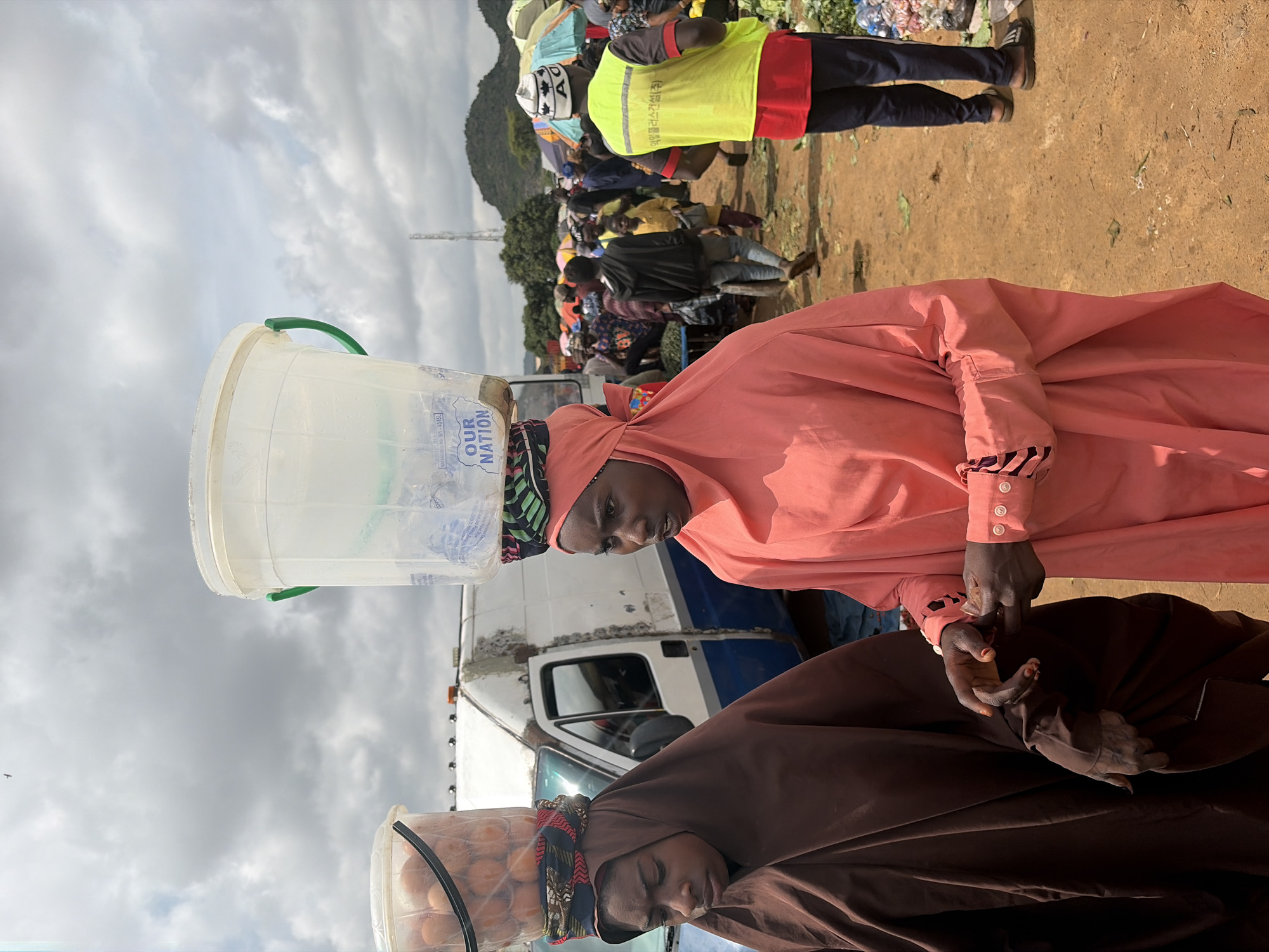 In Gura Top, Nigeria, two young women are seen carrying large plastic buckets of goods on their heads in an outdoor marketplace. The woman on the left, wearing a brown hijab and robe, has a bucket filled with eggs balanced on her head. The woman on the right, wearing a salmon-colored hijab and dress, carries an empty clear plastic bucket with a green handle. They are walking side-by-side, and their hands are near each other, suggesting they might be interacting or walking together. In the background, a white van is parked, and other market-goers can be seen among stalls covered by colorful umbrellas. A man in a yellow vest and dark pants stands to the right, facing away from the camera. The ground is dirt, and the sky is cloudy. The overall scene depicts a busy day at a local Nigerian market.