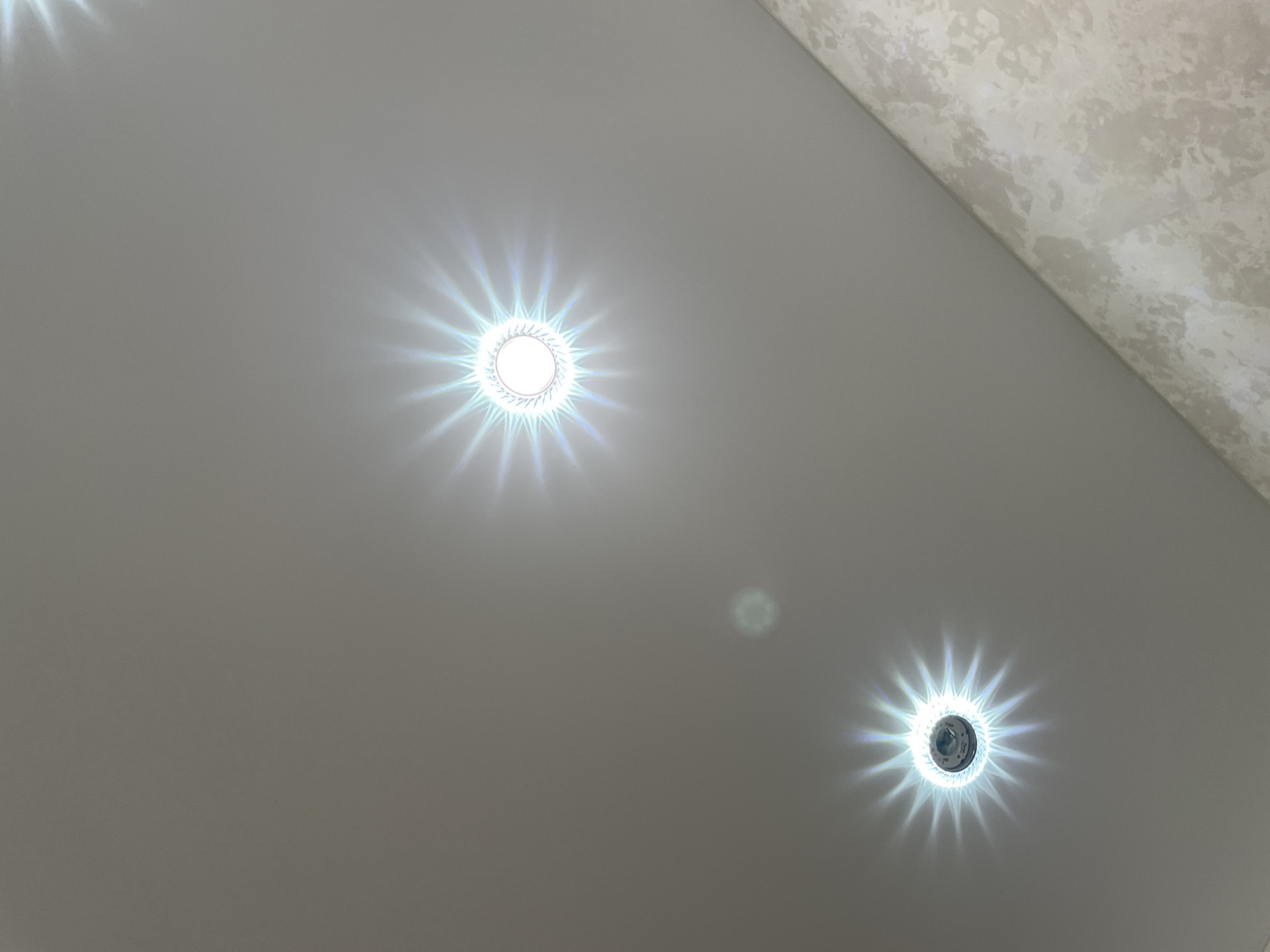 The image shows a ceiling with three recessed lights. Two of the lights are clearly visible and illuminated, casting starburst patterns of light. The third light is partially visible in the upper right corner and appears to be off. The ceiling is a light gray color, and a portion of a textured wall in a cream and beige color is visible in the lower right corner. There are no people or any other objects in the image. The location of this scene in Stavropol, Russia, cannot be determined from the visual information provided.