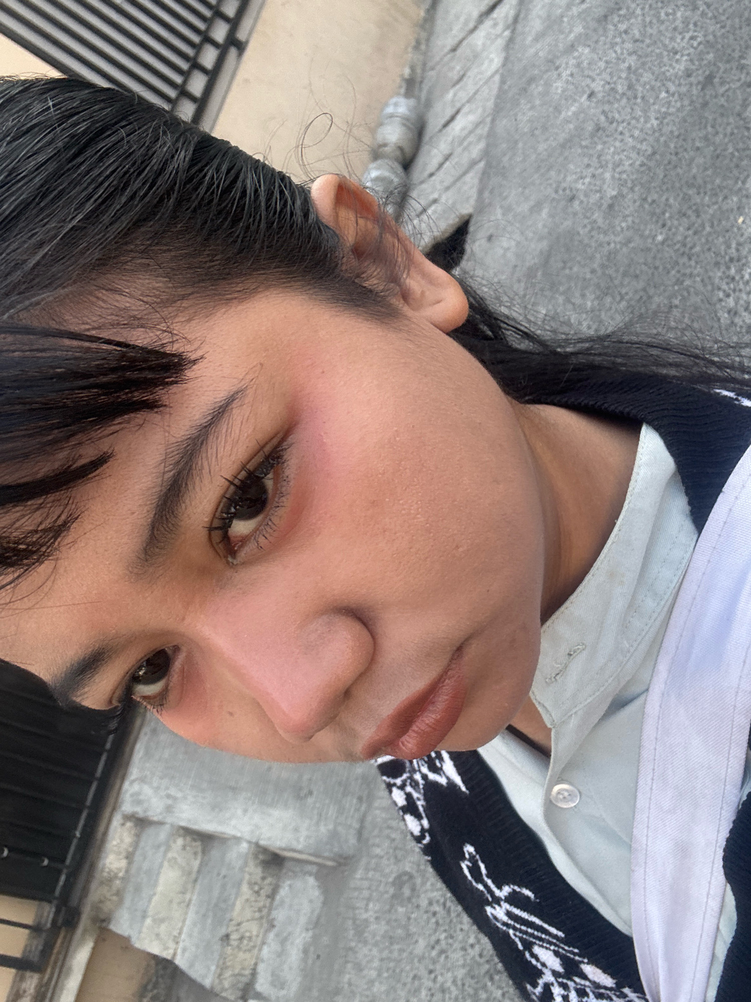 A young person with dark hair and fair skin is captured in a close-up, angled selfie outdoors. They are wearing a light blue button-up shirt with a black and white patterned cardigan over it, and a white strap is visible. The person's makeup includes mascara and lip color. The background shows a textured, grey concrete surface, likely a sidewalk or street, with some vertical elements that could be a fence or railing. The lighting suggests it is daytime. The location is specified as North Jakarta, Indonesia, implying an urban environment.