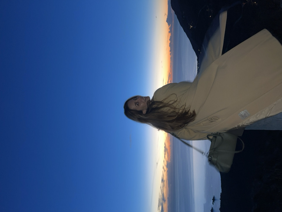 A woman poses in a tan coat against a backdrop of a vibrant blue sky transitioning into a gradient sunset with hues of orange and pink. The horizon shows a landscape of water meeting land. She has long brown hair and smiles subtly. A purse dangles from her arm. Dark, shadowed areas are visible at the edges of the frame, possibly indicating the setting sun or a nearby object.