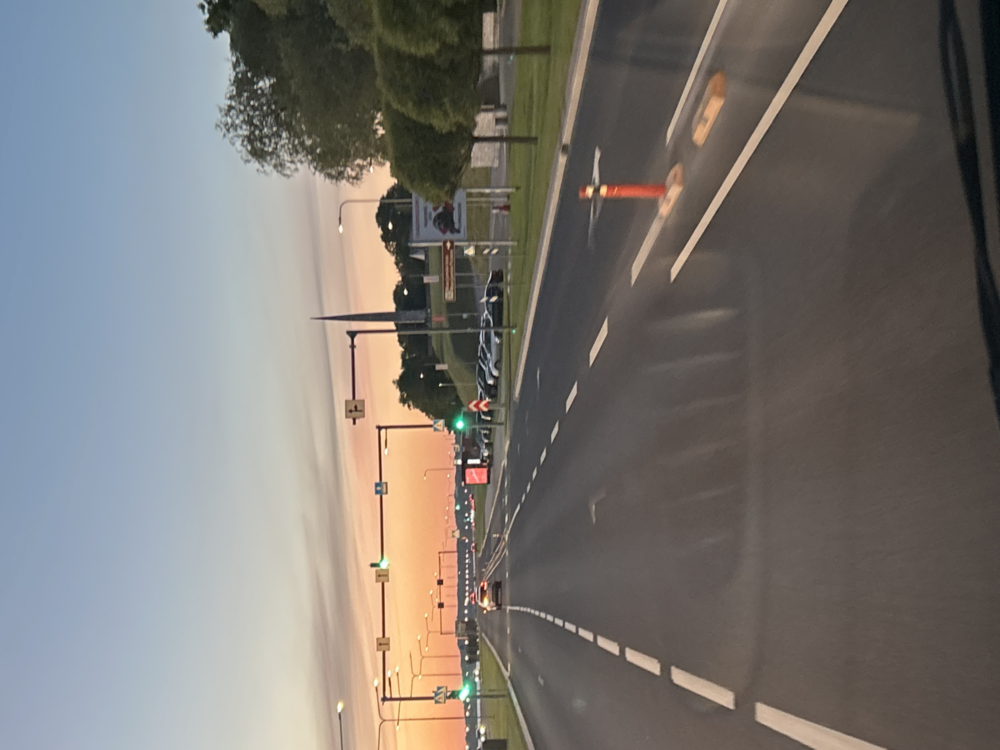 The image depicts a road at dusk in Tallinn, Estonia. The sky is a gradient of pale blue to orange, indicating sunset or sunrise. A multi-lane road stretches into the distance, with white dashed lines marking the lanes. Traffic lights are visible, with some displaying green signals. Several cars are on the road, with one white SUV in the foreground, and other vehicles parked on the side. Trees line the right side of the road, with some appearing to be ornamental topiary. A tall, slender spire, possibly part of a building or monument, is visible in the background. There is also a billboard or advertisement on the right side of the road. A red traffic cone with reflective elements is positioned on the road. The overall impression is of an urban or suburban road scene during twilight.