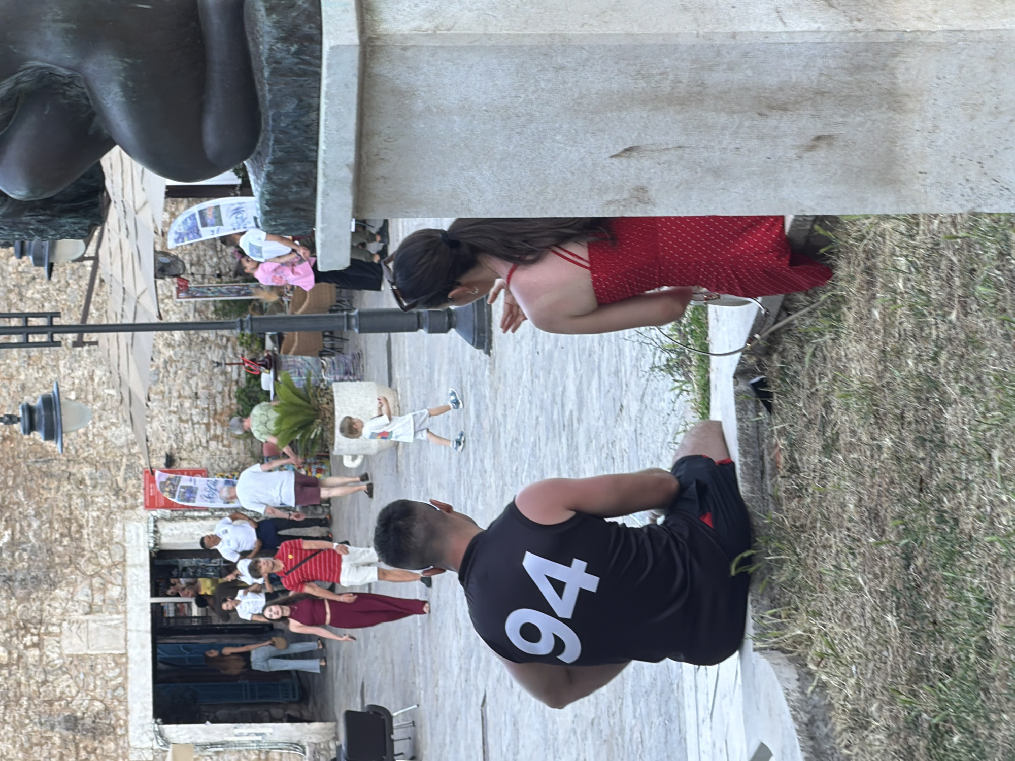In Budva, Montenegro, a group of people are seen in an outdoor urban setting, likely a town square or promenade, adjacent to a stone building with an open entranceway. In the foreground, a man wearing a black tank top with the number "94" in white, and black shorts, sits on a stone ledge facing away from the camera. To his right, a woman in a red, patterned sundress is also seated on the ledge, facing the man and slightly to his right.
Further in the background, several individuals are walking. A young boy in a white t-shirt and grey shorts is walking from left to right. A couple in the mid-ground, a woman in a burgundy outfit and a man in a red and white striped shirt with white shorts, are walking towards the open doorway. Other people are seen in the background near the building, some appear to be shopkeepers or staff members. The environment includes stone paving, grass, and some vegetation. A large stone sculpture is partially visible on the right side of the frame.