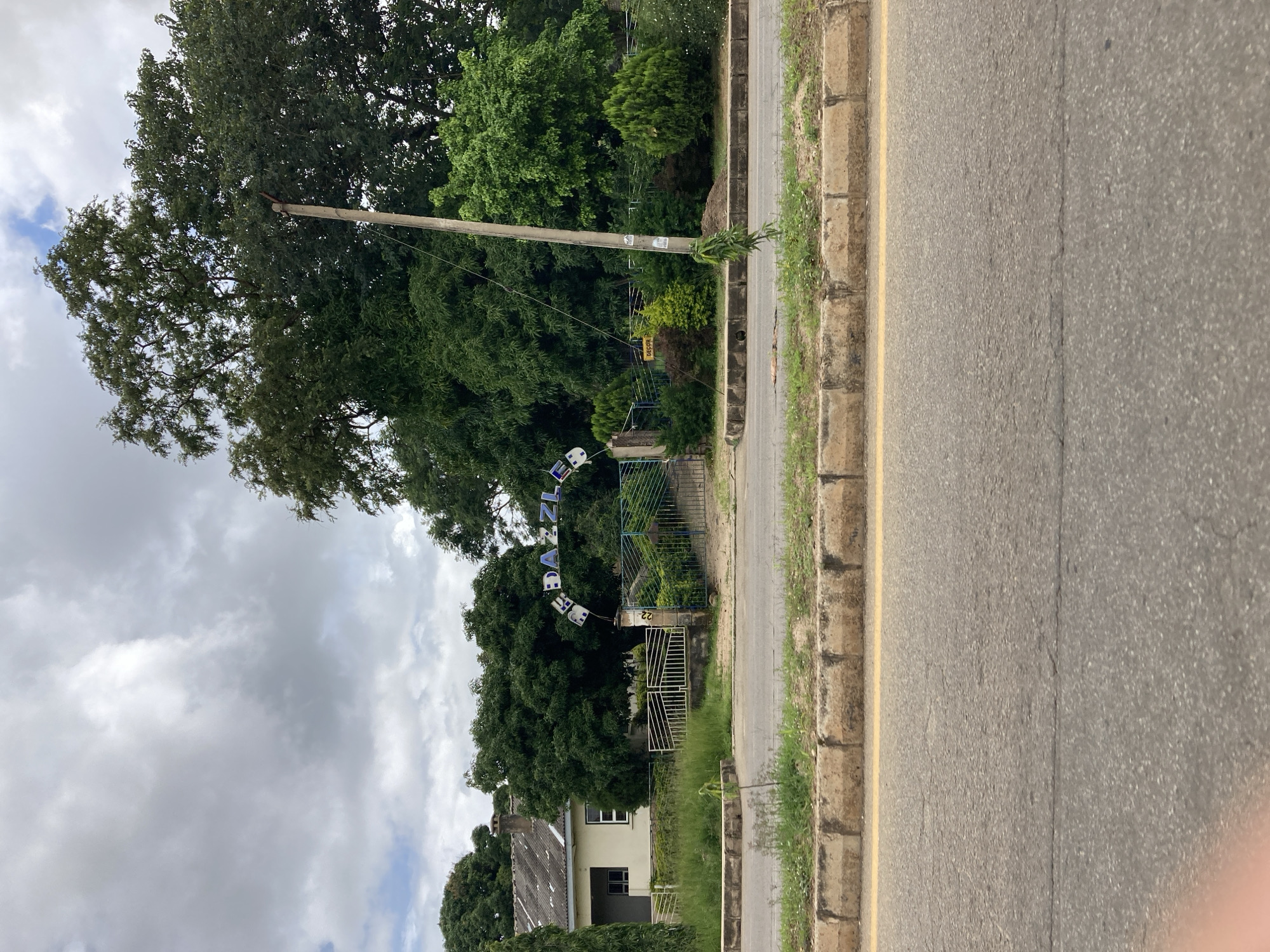 The image captures a street view in Jos, Nigeria. A paved road with a yellow line marking the lane runs across the foreground. A raised curb with worn concrete blocks separates the road from a grassy embankment. Beyond the embankment, a building with a light-colored facade and a dark-tiled roof is visible to the left. In the center, an arched gateway with white metal bars is adorned with blue letters that spell out "RE DA ZZLE." The gateway leads into a property with lush green trees and manicured bushes. A utility pole stands to the right of the gateway, with a small "DECOR" sign attached to it. The sky is overcast with thick, grey clouds.