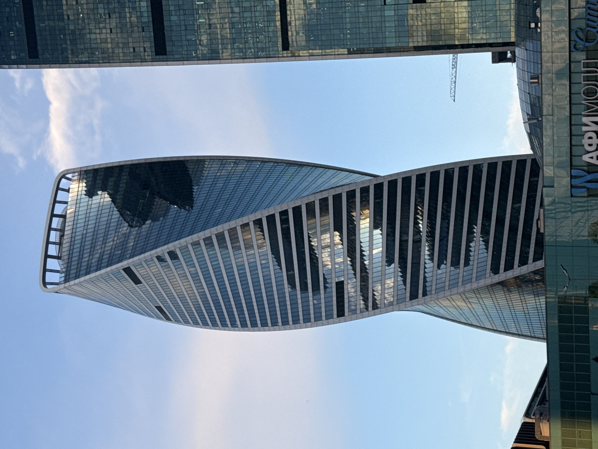 The image shows a close-up, low-angle view of the futuristic Neva Towers complex in Moscow, Russia. The foreground is dominated by the striking, twisting glass facade of the taller Neva Tower 2, which exhibits a helical design. The building's reflective surface mirrors the sky and surrounding structures, creating a dynamic interplay of light and shadow. To the right, a portion of the adjacent Neva Tower 1 is visible, characterized by its more conventional rectangular glass exterior. In the lower portion of the image, a modern glass-covered structure with visible signage that appears to read "АФИМОЛЛ Сити" (AFIMALL City) is present, suggesting the presence of a shopping mall or mixed-use complex. The sky is a clear blue with scattered white clouds, and a faint construction crane can be seen in the distance between the two towers. The scene captures the architectural modernity of Moscow's business district.