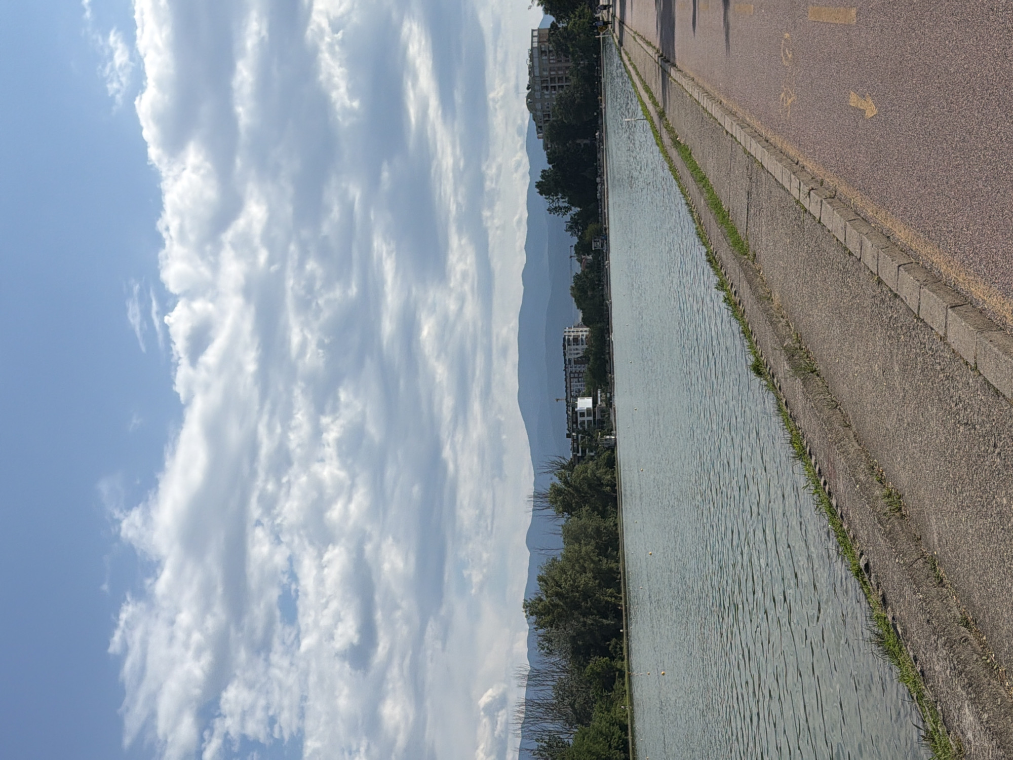 The image depicts a scenic view of the Rowing Canal in Plovdiv, Bulgaria, under a cloudy sky. In the foreground, a paved pathway runs along the edge of the canal, marked with a bicycle symbol and directional arrows. The canal itself is wide and calm, with a few yellow buoys floating on its surface. Across the water, a dense line of trees borders the distant shore, beyond which lie buildings and mountains. The sky is a mix of blue and white clouds, with some areas appearing overcast. A few people are visible walking on the pathway in the distance. The overall scene conveys a peaceful and recreational atmosphere.