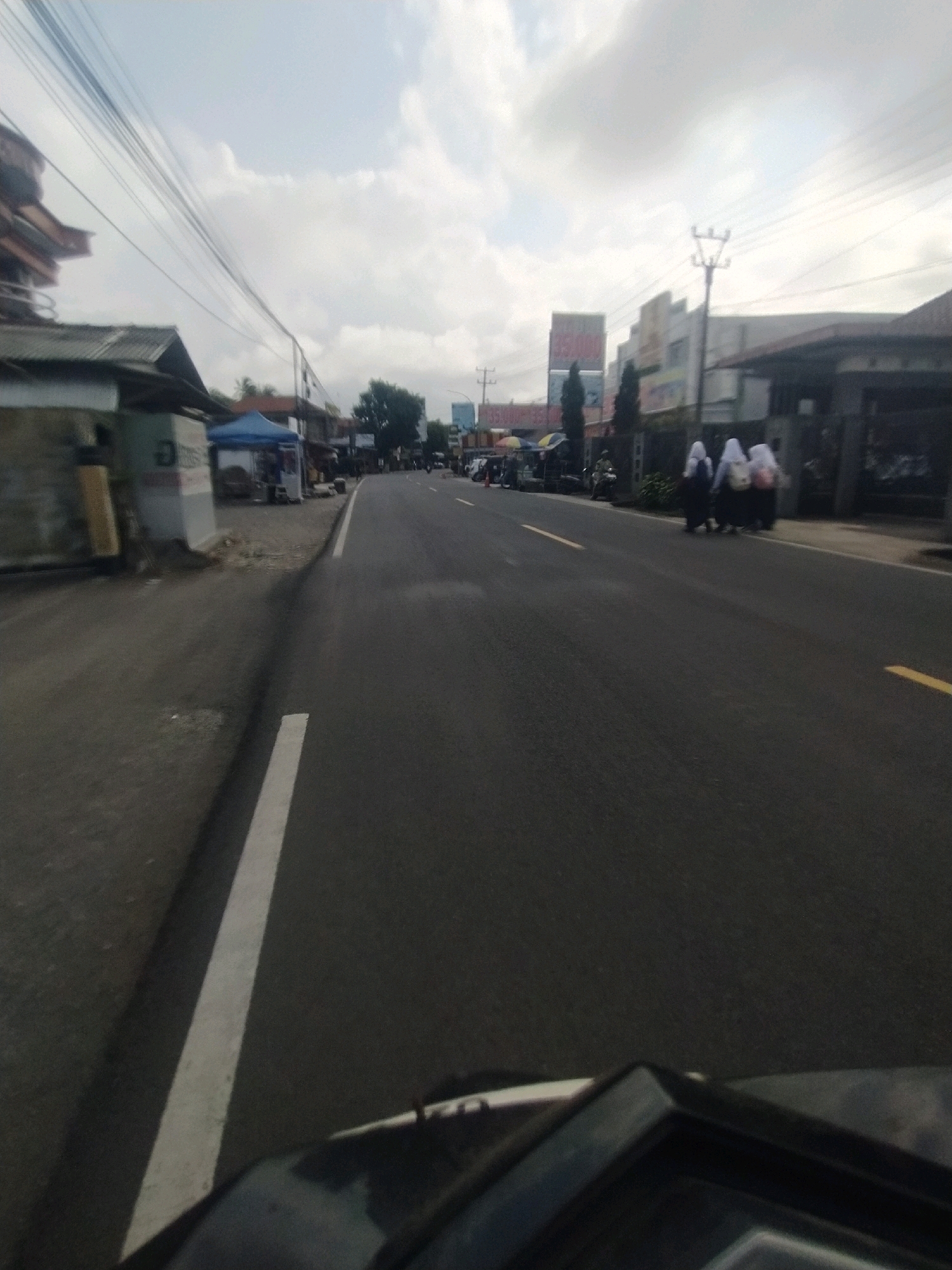The image depicts a street scene in Desa Karangbenda, Indonesia, viewed from the perspective of a person on a motorcycle. A paved road runs through the center of the frame, with a white line marking the lane on the left and yellow lines on the right. Along the left side of the road, there are buildings with corrugated metal roofs, a blue tent, and some market stalls. On the right side of the road, a sidewalk is visible, with a few individuals, possibly students in hijabs, walking away from the camera. Beyond the sidewalk, there are more buildings, including multi-story structures with signs, and trees. Utility poles and wires are present throughout the scene. The sky is partly cloudy, and the overall lighting suggests it is daytime. The foreground shows a portion of the motorcycle, including its handlebars and dashboard.