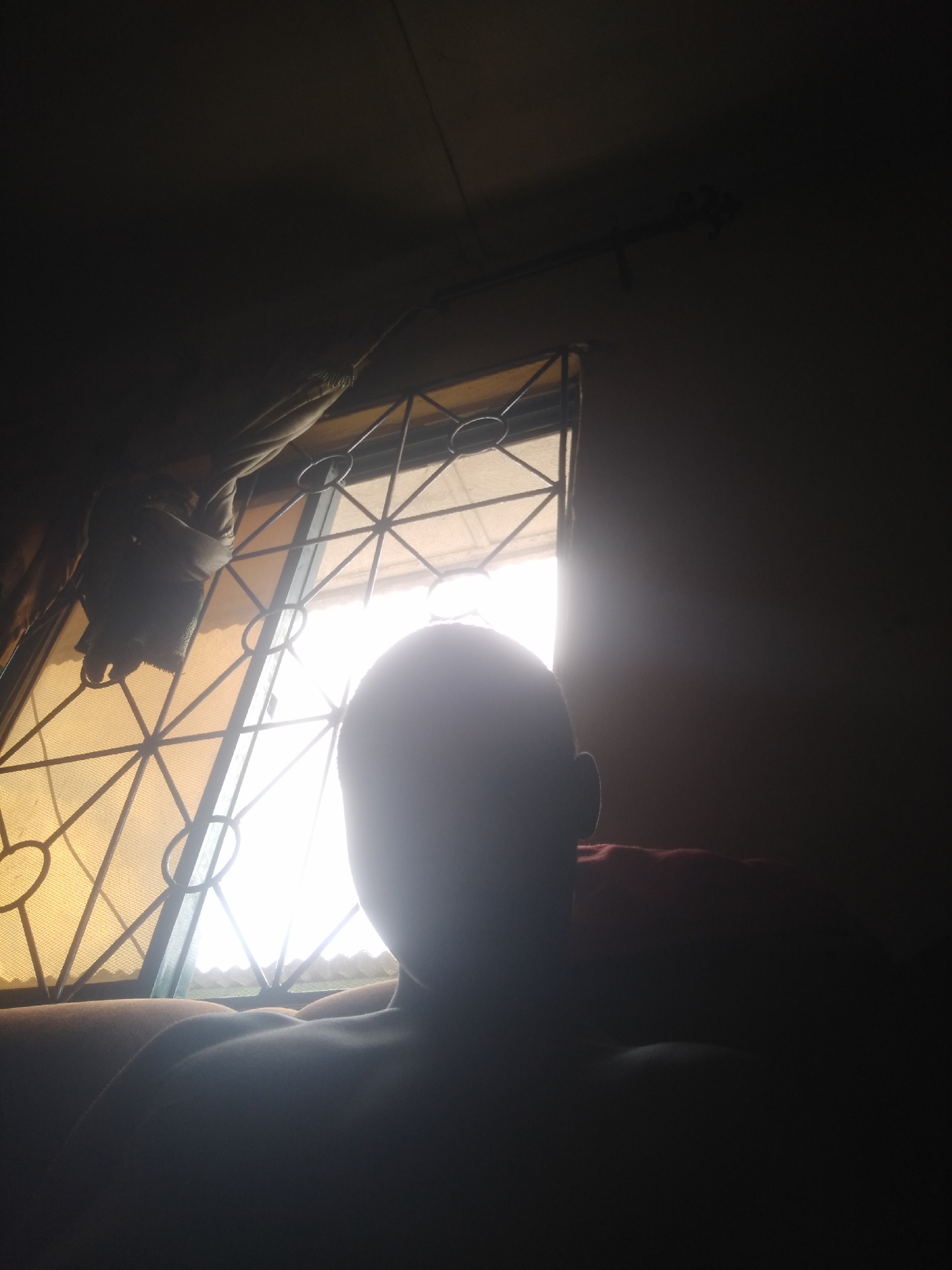 The media file presents an indoor scene with a strong silhouette of a person. The individual, seen from behind or the side, has their head and upper torso visible, completely darkened against the bright light emanating from a window. The room's interior is significantly underexposed, appearing very dark.
The primary light source is a window featuring a prominent metal security grill with a distinct geometric pattern of intersecting bars and circular ornaments. A curtain, possibly light brown or beige, is partially visible on the left side of the window frame. The bright light streaming through the window has a warm, yellowish tint. A hint of red fabric, possibly a pillow or clothing item, is visible near the person's shoulder/head on the right side of the frame.
The image captures a moment during the daytime, likely under sunny conditions, given the intensity of the light from outside. The extreme backlighting creates a high-contrast composition, obscuring facial features and specific details of the room. The architectural detail of the security grill provides a subtle contextual cue for the setting in Shangisha, Nigeria. No visible text is present.