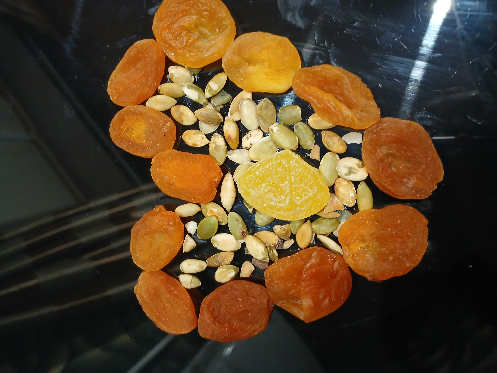 The image shows a collection of dried apricots, pumpkin seeds, and a piece of candied fruit arranged on a glossy black surface. The apricots are various shades of orange and appear slightly wrinkled, indicating they are dried. The pumpkin seeds are a mix of off-white and green, suggesting they are hulled and possibly roasted. The candied fruit is a yellow hue and has a slightly crystalline appearance. The surface reflects light, giving it a polished look. Based on the request's context, this image is presumed to originate from Kamianske, Ukraine.