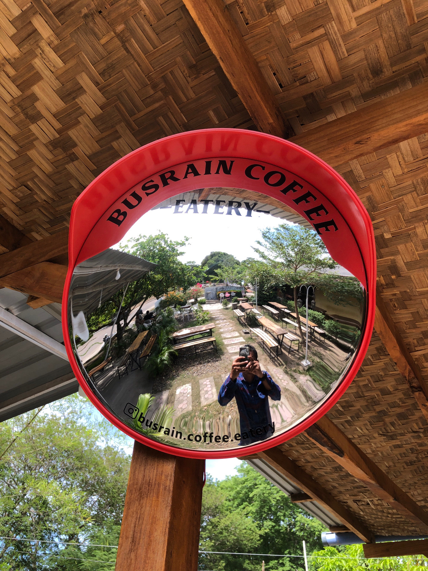 The image shows a convex mirror mounted on a wooden post. The mirror reflects an outdoor seating area of a cafe. The cafe has wooden tables and benches set up on a stone-paved ground, surrounded by greenery and trees. The roof structure above the seating area is made of woven bamboo. The mirror has "BUSRAIN COFFEE EATERY" written around its rim. Below the text, an Instagram handle "@busrain.coffee.eatery" is visible. In the reflection, a person wearing a blue shirt is taking a selfie with a camera. The location is identified as Tambonbaroh, Indonesia.