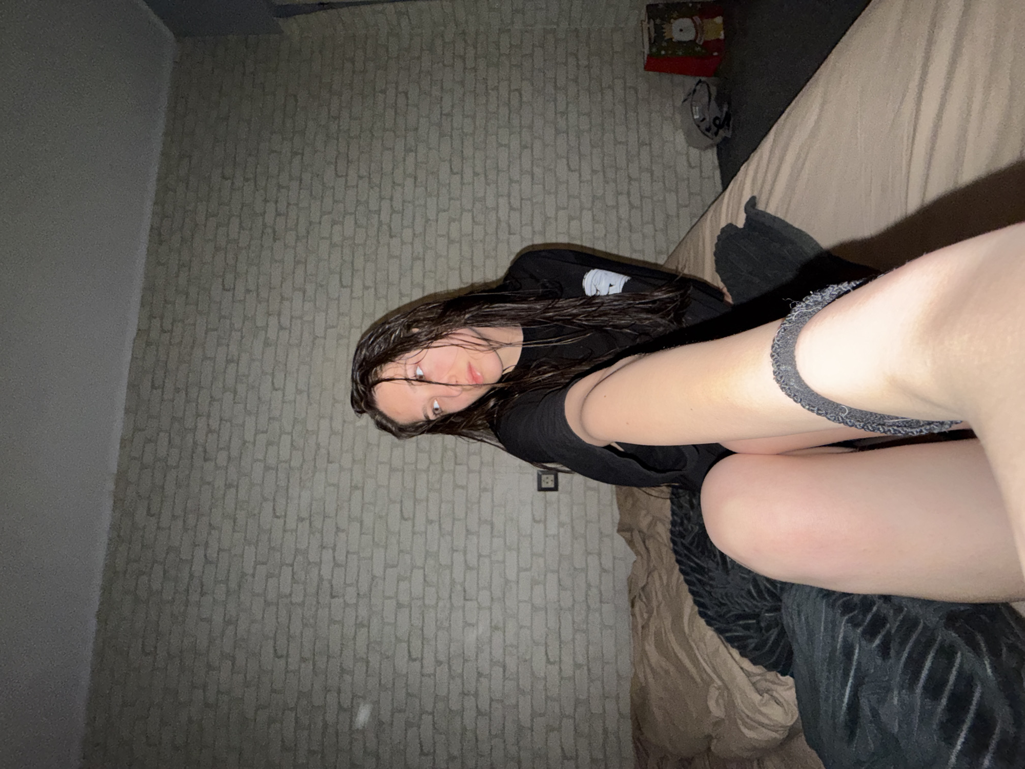 A young woman with wet, dark hair is lying on a bed. She is wearing a black t-shirt and is positioned diagonally in the frame. The room has a brick-patterned wallpaper. The bed has beige sheets, black blankets, and a gift bag visible in the corner. An electrical outlet can be seen on the wall. It is impossible to determine the location in Sofia, Bulgaria.