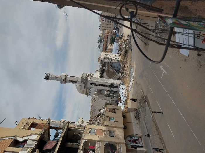 This image depicts a severely damaged urban landscape at an intersection in Khan Yunis, Palestinian Territory, viewed from an elevated vantage point.
The central feature is a prominent white mosque with a tall minaret and a dome, which has sustained extensive structural damage, with large sections of its walls collapsed, revealing internal rubble. Surrounding the mosque are vast piles of debris and building remnants.
To the immediate left, a multi-story building exhibits partial collapse, exposed interiors across multiple floors, damaged balconies, and laundry items hanging from remaining structures. A poster featuring "SQUID GAME" is visible on its ground-level facade. In the background, numerous other multi-story buildings also show signs of significant damage or partial collapse.
The street below is a paved road with a single white directional arrow painted on its surface. The road is flanked by dirt shoulders and additional piles of rubble. At least five individuals are present: one person walks on the left carrying a dark object, another stands centrally in the street, a third crouches near the left sidewalk, and two others are visible further in the distance.
In the immediate right foreground, a dark metal balcony railing with a piece of striped fabric draped over it partially obstructs the view, indicating the elevated perspective from which the photograph was taken. The sky above is partially cloudy.