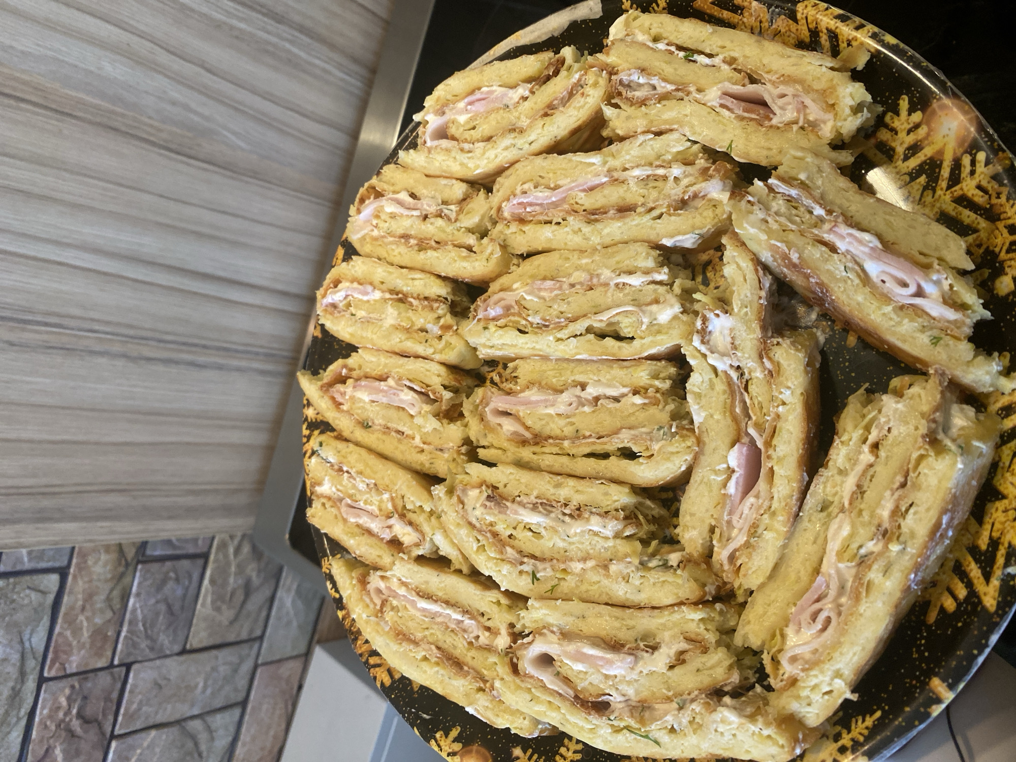 The image displays a close-up, high-angle view of a large, dark serving platter filled with numerous slices of savory rolled food, likely prepared for a meal or gathering. The items are arranged in rows, almost covering the entire platter.
Each slice reveals multiple layers: an outer, light golden-yellow, soft-looking dough (resembling a pancake or crêpe), wrapped around a filling. The filling consists of a generous spread of white, creamy substance, possibly cream cheese or a mayonnaise-based mixture, with visible green specks suggesting herbs like dill or parsley. Layered within the creamy filling are thin, pinkish slices of what appears to be ham or another cold cut. The rolls have been cut into thick, oval-shaped slices, showcasing their intricate spiral structure.
The platter itself is dark-colored and features decorative gold or yellow patterns, resembling snowflakes or starbursts, particularly visible along the edges and in gaps between the food items.
In the background, to the left of the platter, a wooden surface with a distinct grain pattern is visible, likely a countertop or table. Below this, on the bottom left, a section of floor with light-toned, geometric tile patterns can be seen.
The scene suggests an indoor setting, possibly a kitchen or dining area, in Sliven, Bulgaria, where these prepared appetizers or finger foods are ready to be served or consumed. There is no visible text or identifiable people in the image.