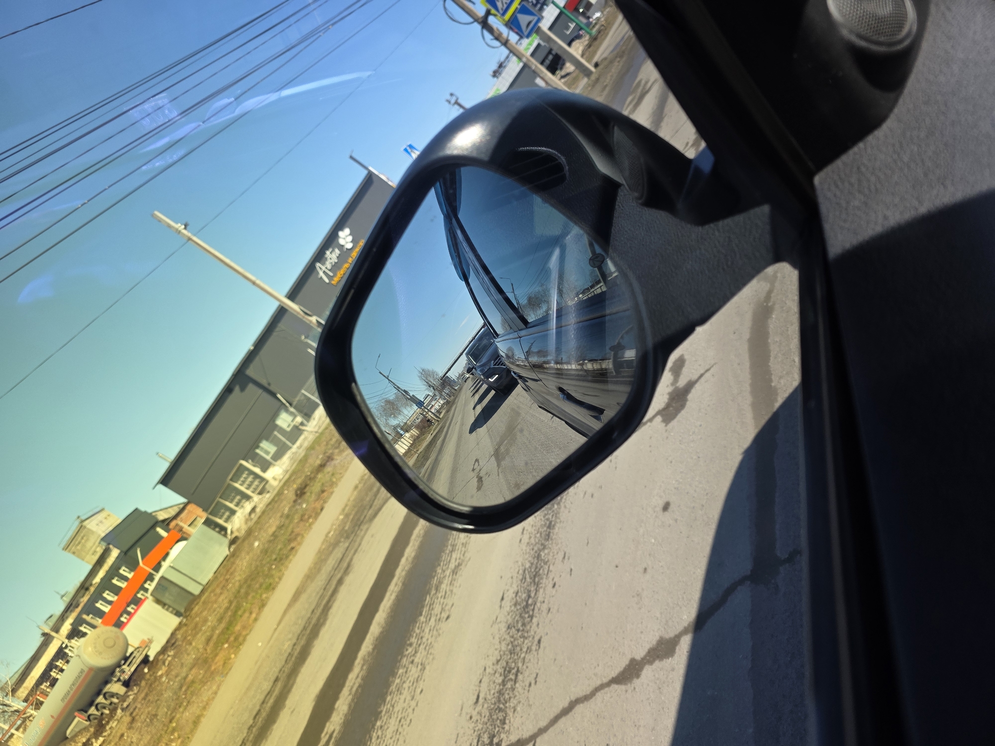 A view from inside a car, focusing on the passenger side mirror. The mirror reflects a street scene, including buildings, other vehicles, and overhead wires. A large building with the name "Astra" is partially visible. The location is the city of Chaikovsky, Russia, based on the name "Astra" on the building. The sky is blue, and the day appears sunny. The road surface is gray.