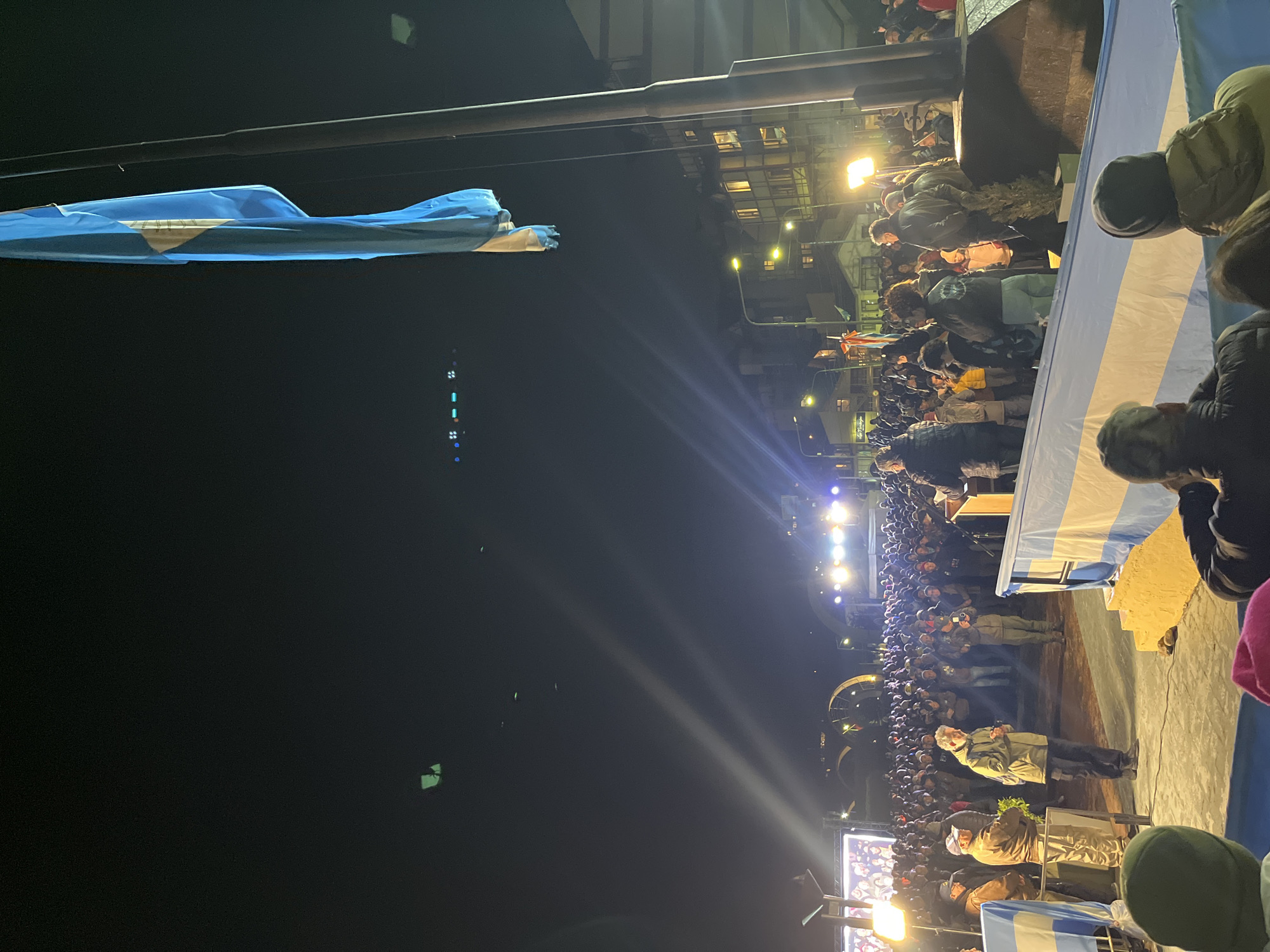 The image captures a nighttime event in what is presumably Ushuaia, Argentina, based on the presence of an Argentinian flag. A crowd of people is gathered in front of a raised platform or stage, which is draped with an Argentinian flag. The stage has a podium, and several individuals are standing on it. Bright lights illuminate the stage and the surrounding area, making it difficult to discern specific details about the people in the crowd. The background features a building illuminated by streetlights, indicating an urban setting. A flag is visible, possibly the Argentinian flag, hanging from a pole on the left side of the frame. The overall atmosphere suggests an outdoor gathering, possibly a ceremony or event with a significant crowd.