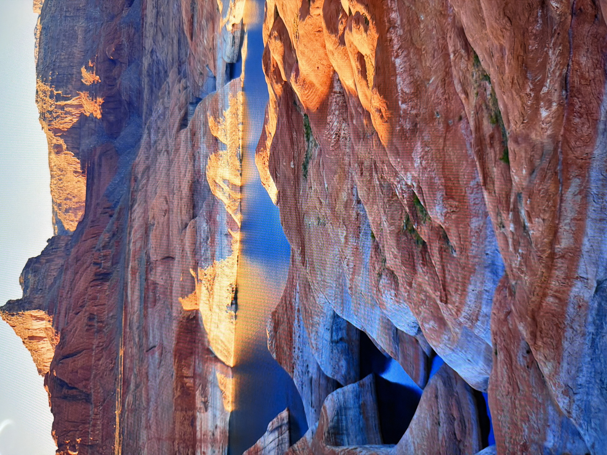 The image shows a panoramic view of a desert landscape with a body of water. The terrain is characterized by layers of red and white rock formations, likely sandstone, sculpted by erosion into intricate shapes. The water, a deep blue, reflects the sky and the surrounding rocks, creating a striking contrast. In the background, towering mountains with sharp peaks rise against a clear, light blue sky. The sunlight casts long shadows and highlights the textured surfaces of the rocks, indicating it is either early morning or late afternoon. There are no people or man-made objects visible in the image. This landscape is characteristic of desert regions such as the American Southwest, not Kyiv, Ukraine.