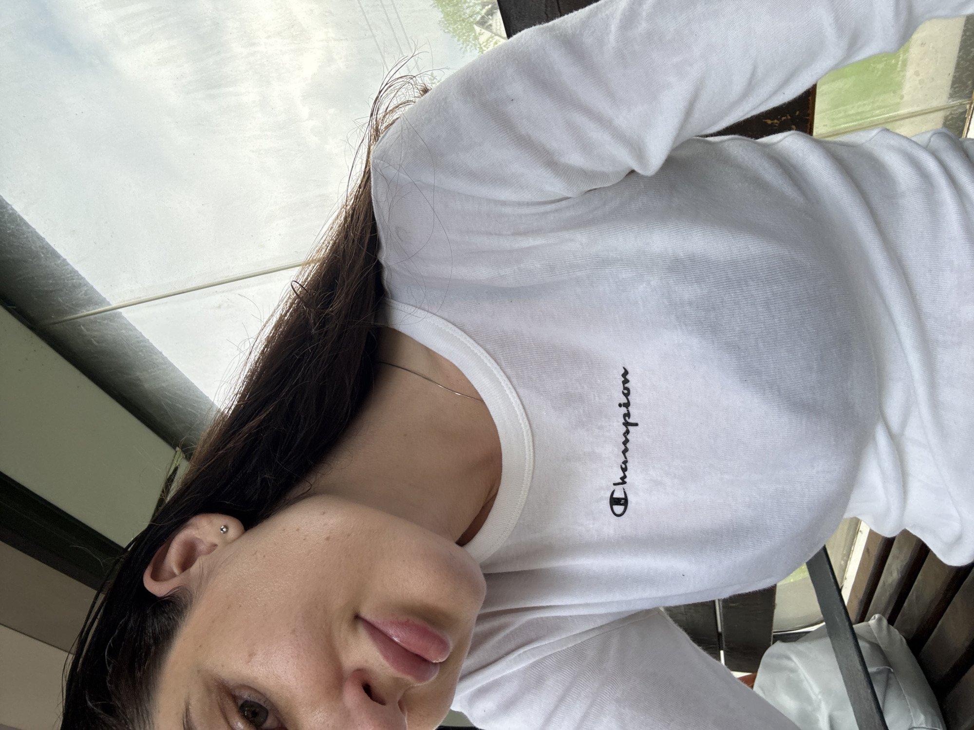 A person wearing a white "Champion" long-sleeved shirt poses indoors, with part of a window and a wooden bench visible. The location is in the city of Unknown, Unknown. The individual has dark hair and an ear piercing. A silver necklace is visible at their neck. The person's face, including an eye and their lips, is partially visible. The window shows a cloudy sky. The image appears to be taken from a close-up, low-angle perspective.