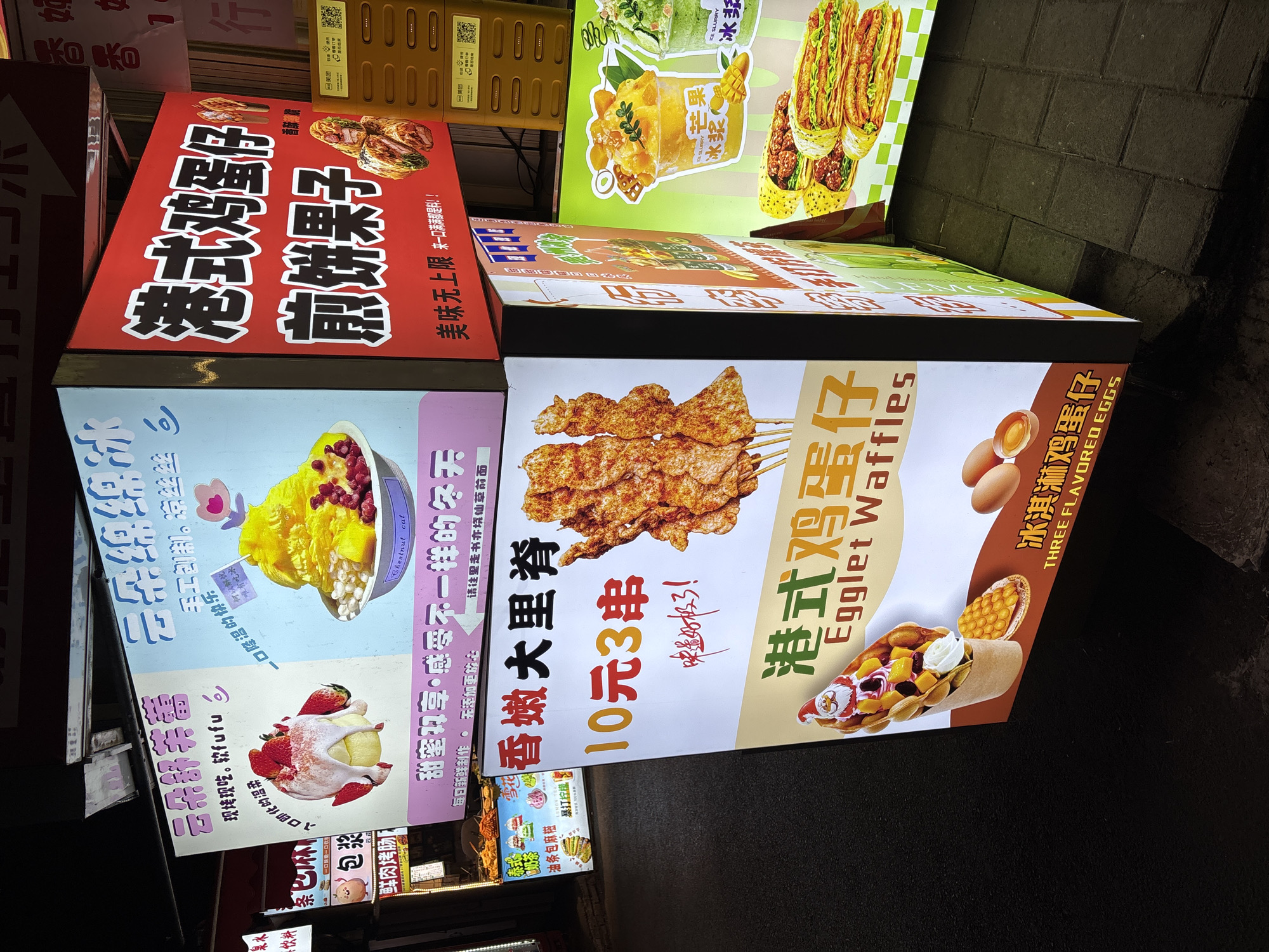 The image shows an outdoor display of illuminated food advertisements, likely in a commercial district. Several rectangular lightboxes are stacked and arranged close together, advertising different street foods. The advertisements include items like grilled skewers of seasoned pork, Hong Kong-style egg waffles, various dessert bowls with mango, and savory pancakes with meat. The characters appear to be Chinese, indicating a location in China, with "Nan'an District" mentioned in the surrounding text. The environment is dimly lit, suggesting it is either evening or nighttime. The sidewalk in the foreground is paved with dark tiles. Some of the signs have translated English text, which indicates a desire to appeal to a broader audience. The signs are primarily red, blue, yellow, and green. The photograph has a slight upward angle, looking at the illuminated food advertisements.