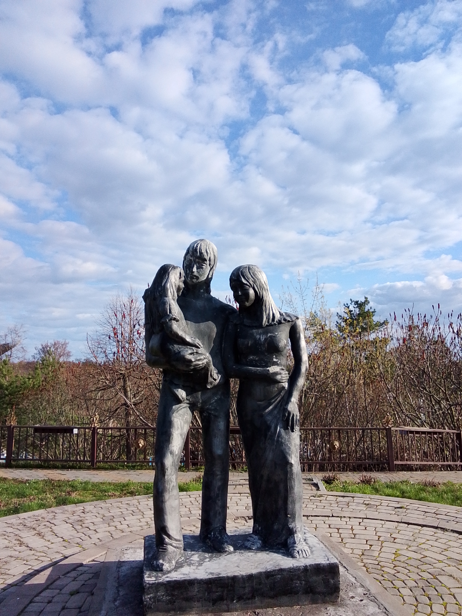 This image captures a bronze or dark metal statue of a family group, set outdoors in a park or public garden in Khlevnoye, Russia. The statue features three figures: a man holding a young child, and a woman standing beside them. The man, depicted with short hair and casual attire, cradles the child in his left arm, looking down at them. The child leans against the man, facing away from the viewer. The woman, with long hair and a draped garment, stands to the man's right, gazing gently towards him and the child. The poses convey a sense of family unity, tenderness, and protection.
The statue rests on a circular paved area made of light-colored cobblestones, surrounded by green grass. Distinct shadows cast by the figures indicate bright daylight, suggesting the time is midday or early afternoon. The weather appears fair, with a clear blue sky dotted with scattered white cumulus clouds. In the background, a low metal fence delineates the area, behind which are numerous bare trees and bushes, some with reddish foliage or seed heads, characteristic of late autumn or early spring. A few evergreen trees are also visible further back. There is no visible text on the statue or in the surrounding environment.