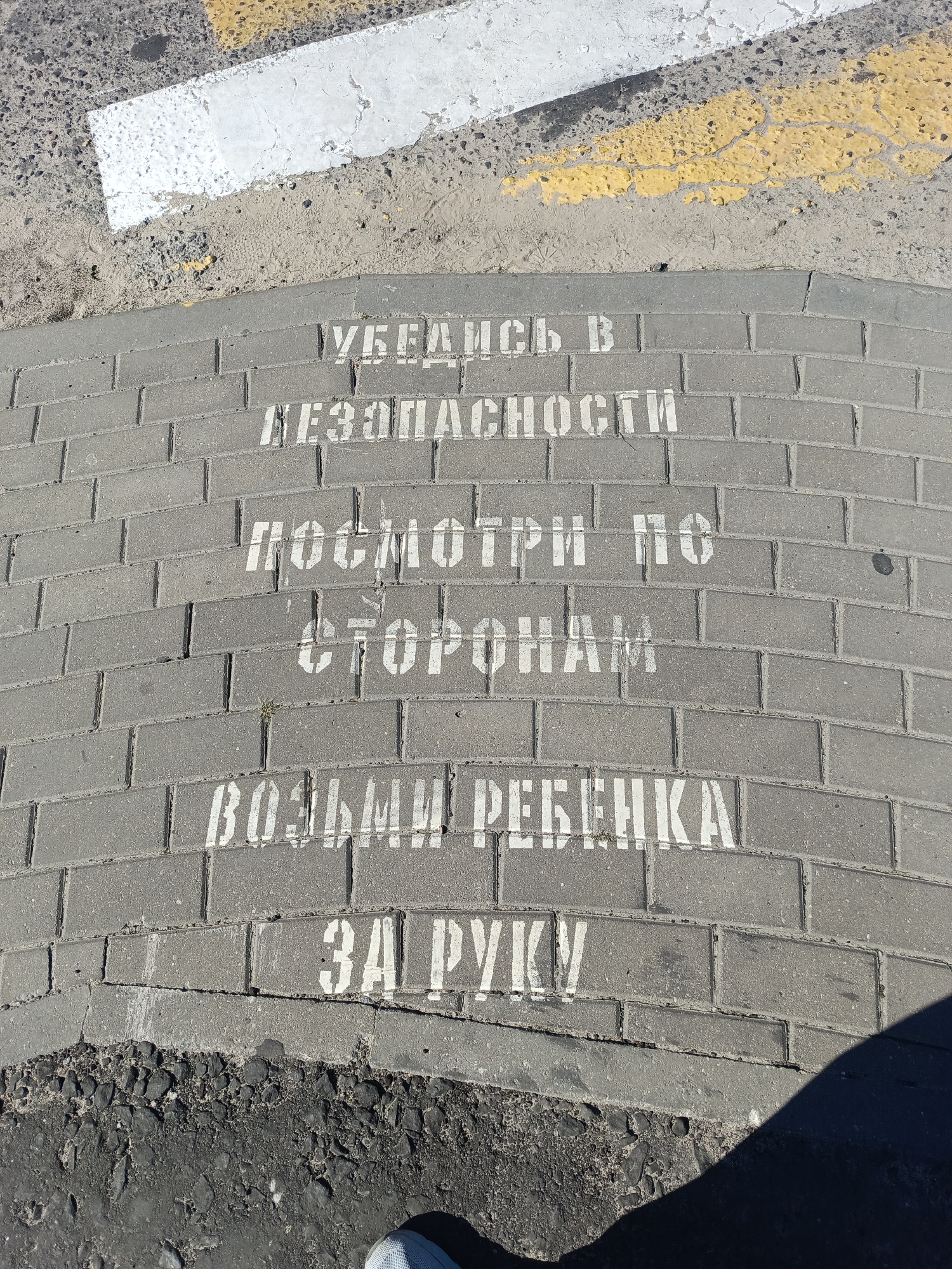 The image shows a section of a sidewalk with white stenciled text painted on it. The text appears to be a safety warning in Russian. The sidewalk is made of grey rectangular pavers, and the text is arranged in multiple lines, curving with the sidewalk. The first line reads "УБЕДИСЬ В" (Be sure of), the second line reads "БЕЗОПАСНОСТИ" (safety), the third line reads "ПОСМОТРИ ПО" (Look to the), the fourth line reads "СТОРОНАМ" (sides), the fifth line reads "ВОЗЬМИ РЕБЕНКА" (take the child), and the last line reads "ЗА РУКУ" (by the hand).
The background of the image shows asphalt and some yellow and white road markings. A shadow of a person is cast on the right side of the image, and a shoe is visible in the bottom center. The overall scene suggests an outdoor urban environment, likely near a road crossing, where parents are being reminded to ensure their child's safety before crossing.
This specific text, "Убедись в безопасности, посмотри по сторонам, возьми ребенка за руку," is a common safety reminder for pedestrians, especially when crossing roads with children, in Belarus. While the image itself does not explicitly state the location as Ivatsevichy, Belarus, this type of public safety messaging is prevalent in Belarusian cities.