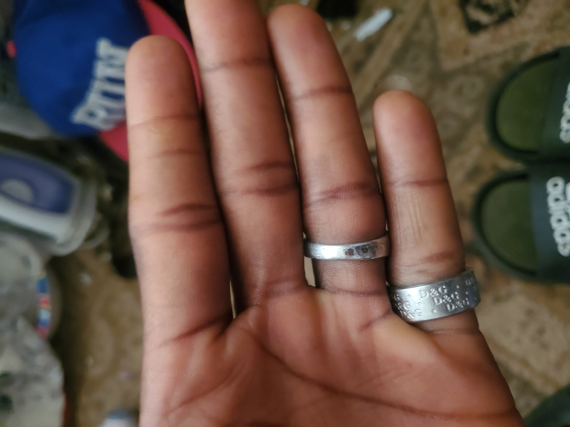 A close-up, eye-level shot shows a person's left hand with three rings on their fingers. The person's skin tone is dark brown. The rings are silver-colored. The ring on the index finger is wider and has the engraved text "D&G" repeatedly around it. The ring on the middle finger is thin and plain. The ring on the ring finger is also thin and plain. In the background, out of focus, are two green Adidas slides, some brown carpet, and a portion of a blue baseball cap. The lighting is bright, casting minimal shadows. There are no identifiable landmarks or context to determine the location within Jos, Nigeria.