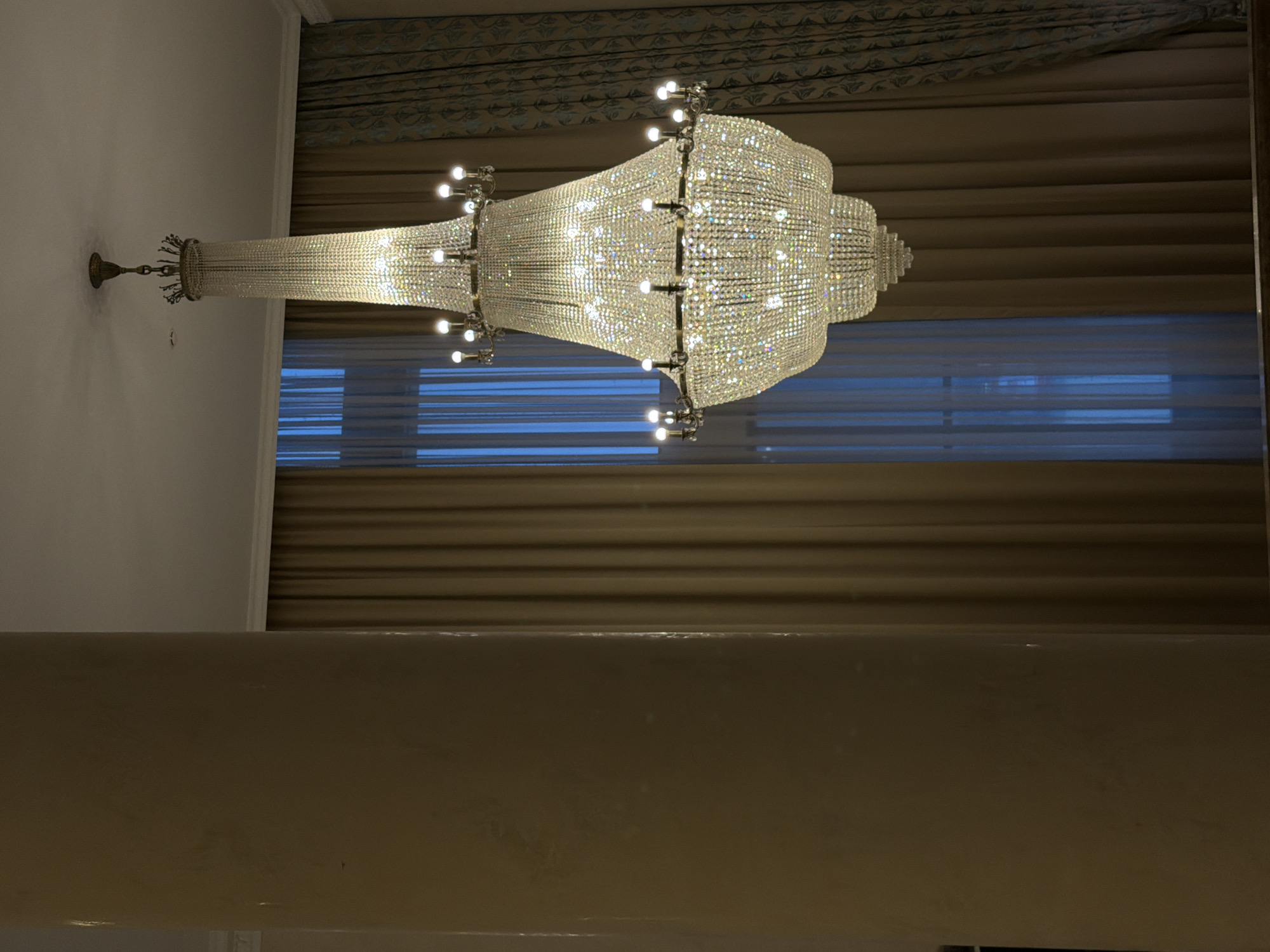 The image shows a large crystal chandelier hanging from the ceiling. The chandelier is multi-tiered and studded with numerous lights. In the background, there are curtains, and a window is visible. The location is not discernible from the image; there is no evidence to indicate that it is in Izhevsk, Russia.