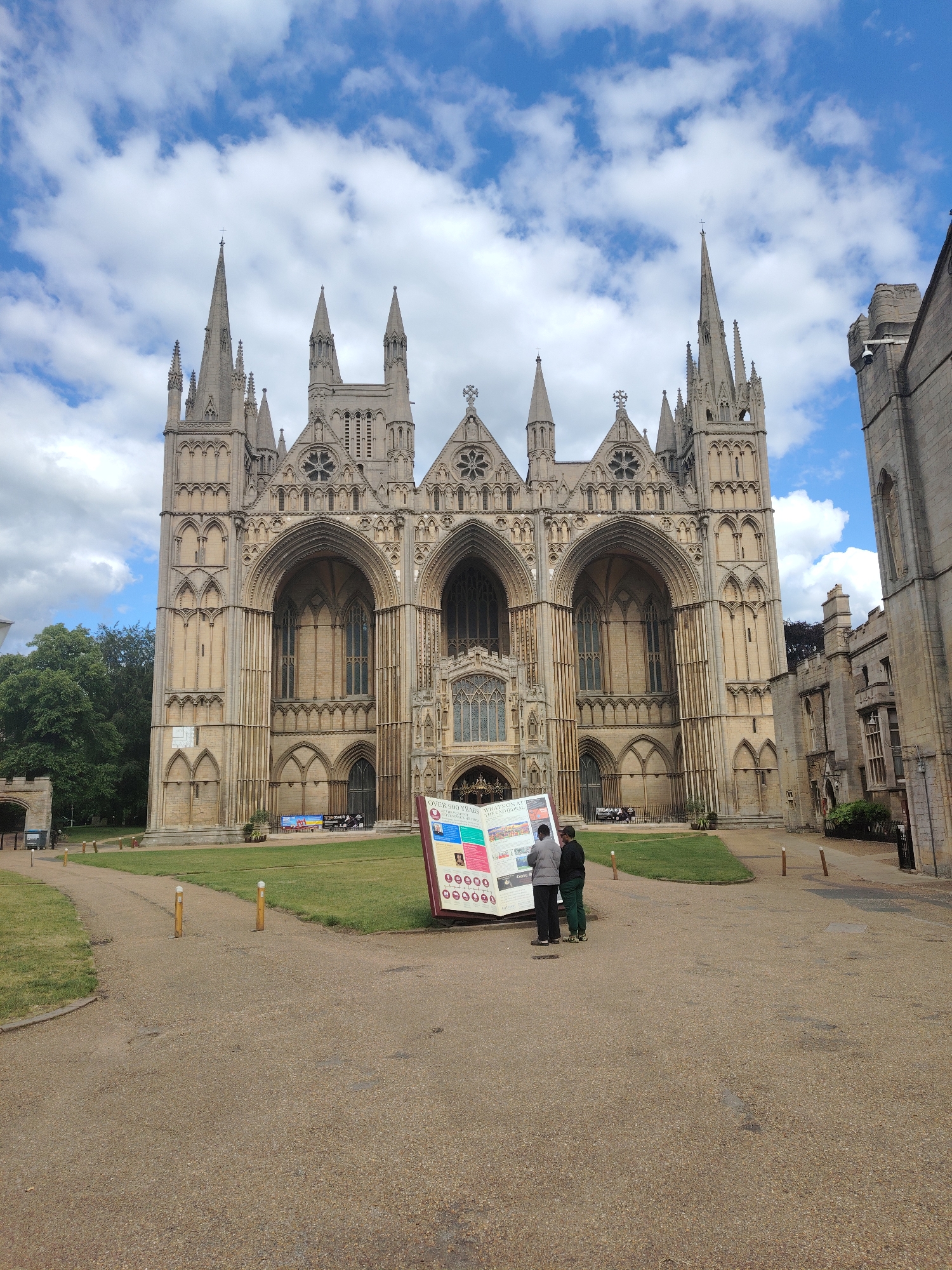 The image depicts the West Front of Peterborough Cathedral in Peterborough, United Kingdom. The cathedral is a large, light-colored stone structure in the English Gothic style, featuring a prominent tripartite facade composed of three towering pointed arches, intricate tracery, rose windows, and numerous spires and turrets, with a central spire being the tallest.
In the foreground, a wide, light brown gravel path extends from the bottom of the frame towards the cathedral. Green grass lawns flank the path on both sides. Several wooden bollards with white tops are spaced along the edges of the gravel path.
Mid-ground, a large, angled informational stand with multiple panels of text and images is positioned on the gravel path. Two individuals stand in front of this sign, facing it, appearing to read. One person wears a light grey jacket and dark trousers, while the other wears a dark top and green trousers.
To the right of the cathedral, a multi-story stone building with windows and battlements is partially visible. On the far left, behind a large green tree, a stone archway or gatehouse can be seen. The sky is bright blue with scattered white cumulus clouds. The scene is illuminated by natural daylight.