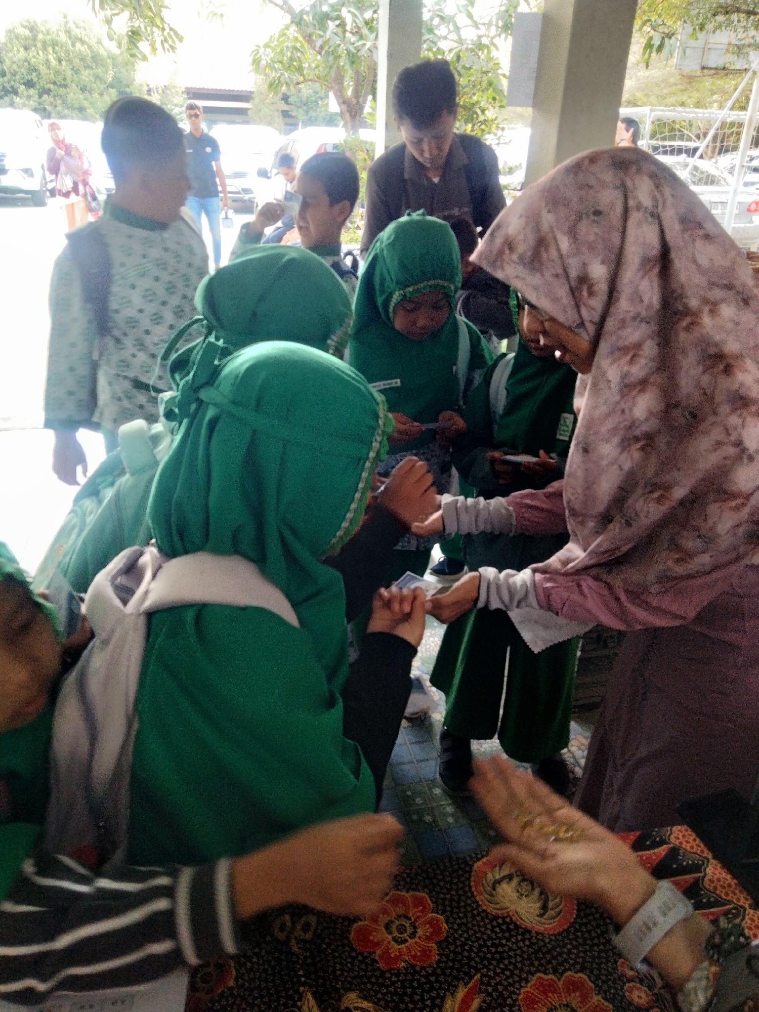 This is a medium-shot, eye-level, full-color photograph taken outdoors in Sanggrahan, Indonesia. The scene depicts a group of people, primarily children wearing green hijabs and uniforms, gathered around what appears to be an informal event or gathering. The background suggests a public space, possibly near a building with a covered walkway, indicated by the presence of pillars and a roofline. Trees and foliage are visible, hinting at a natural setting. Several vehicles, including what looks like white vans, are parked in the background, suggesting the location is accessible by road.
In the foreground, several children are visible, some in focus and others slightly blurred. One child in the center is wearing a green hijab and uniform, looking down. To the left, another child in a green hijab is partially visible. To the right, an adult woman wearing a patterned hijab and a long-sleeved dress is interacting with a child, with their hands positioned as if exchanging something or engaged in a game. The woman's hand is open, showing small objects that might be coins or trinkets. Another adult man in a dark shirt and sunglasses is standing further in the background, looking towards the left. The ground appears to be paved with tiles. The overall atmosphere suggests a community event or a school-related activity.