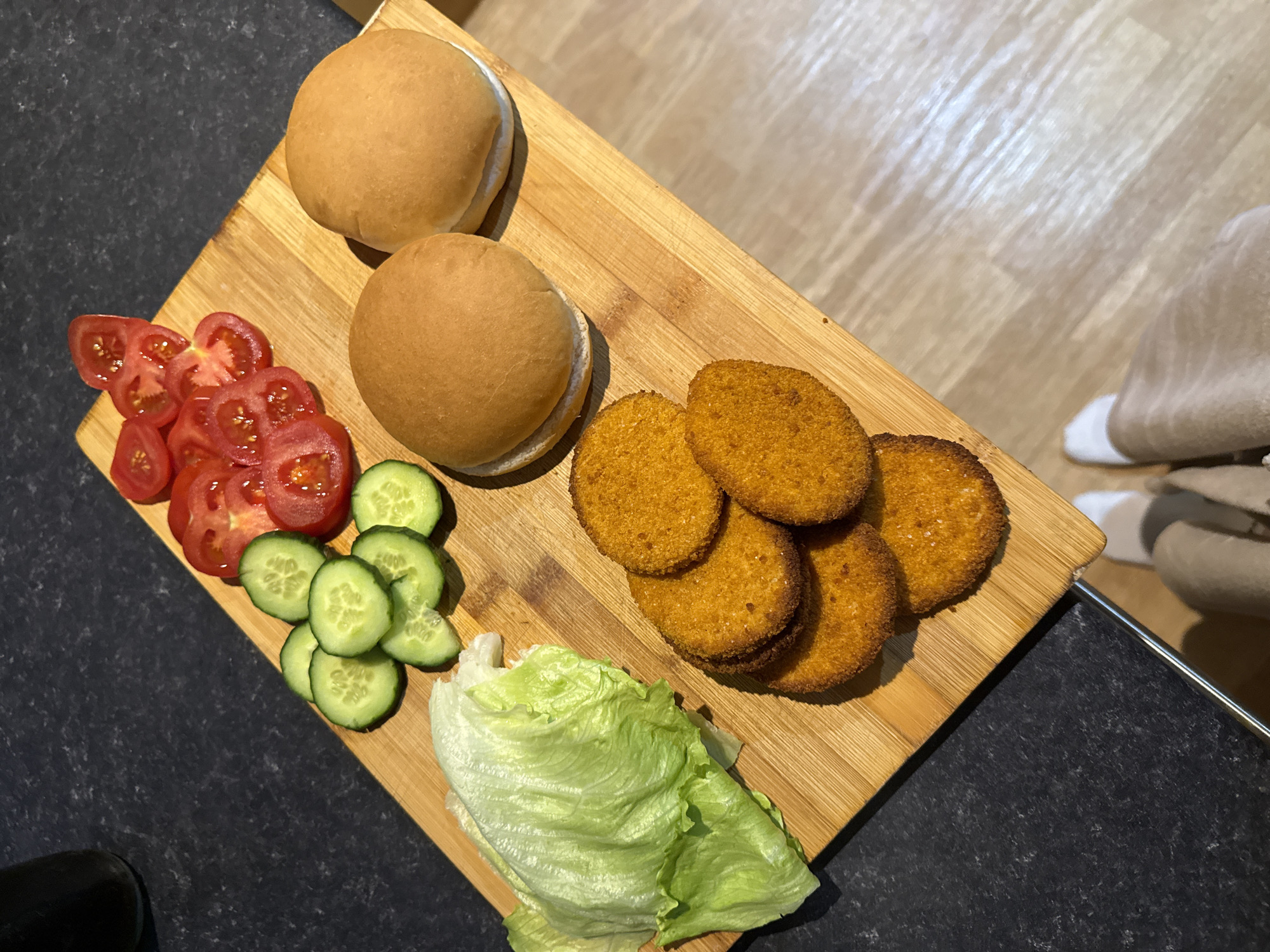 The image displays a high-angle view of food items arranged on a wooden cutting board, indicative of meal preparation within a domestic kitchen setting. The cutting board is positioned diagonally on a dark, speckled countertop. Arranged on the board are various ingredients: a quantity of fresh green lettuce leaves to the left, approximately ten circular slices of green cucumber in the center-left, and a cluster of ten or more sliced red tomatoes in the upper-right. To the immediate right of the vegetables, two light brown burger buns are present, each appearing to be split horizontally. A stack of six to seven circular, breaded, golden-brown patties, consistent with chicken or fish burgers, is situated on the lower-center of the board. A dark, cylindrical container is partially visible on the countertop in the upper-left corner of the frame. The lower legs and feet of an individual, wearing white socks and light-colored trousers or leisurewear, are visible on a light-toned wooden floor in the bottom-right corner of the image. The specific location within Great Yarmouth, United Kingdom, is not discernible from the provided visual evidence; however, the scene is consistent with a private residential kitchen.