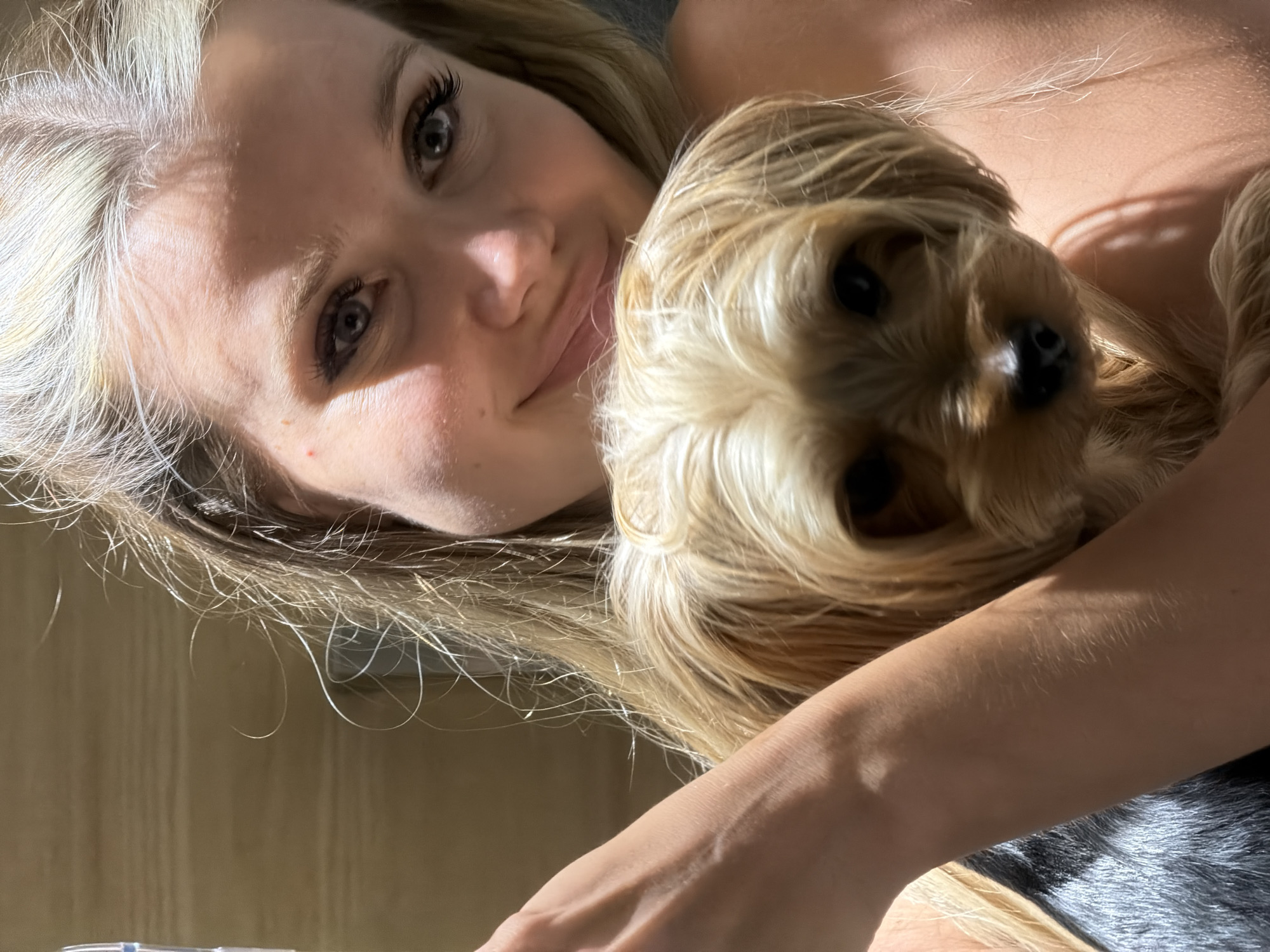 A young blonde woman is holding a small, light brown dog. The woman is smiling and looking at the camera, with her hair falling around her face and shoulders. The dog is nestled in her arms, also looking towards the camera. They are in a dimly lit room with a plain background. The overall impression is one of affection and closeness between the woman and the dog. There is no specific information about the location in Thep Krasatti, Thailand.