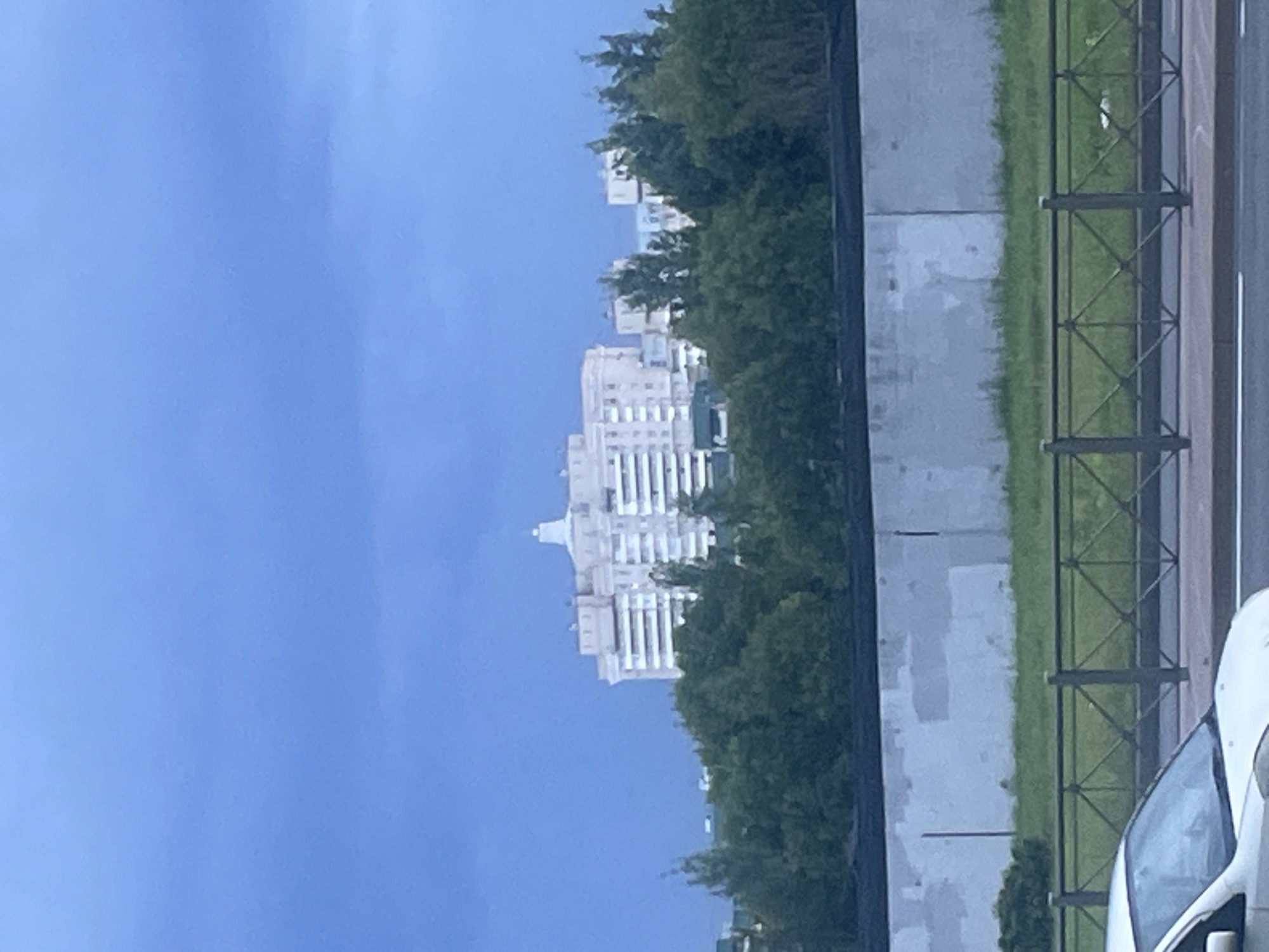An overcast sky, a muted blue, hangs over a residential building and a line of trees in Saint Petersburg, Russia. The building, light-colored with multiple stories and white-framed windows, is positioned on a slight rise, its upper portion adorned with a decorative element that resembles a small dome or spire. Dense, green trees form a natural barrier between the building and the foreground. Below the trees, a concrete wall, weathered and marked with patches of lighter and darker shades, stretches horizontally across the frame. Beyond the wall, a patch of green grass is visible, bordered by a black metal fence with a diamond pattern. In the very bottom of the image, the front section of a white car is visible, parked on a road. The scene suggests a typical urban residential area under a cloudy sky.