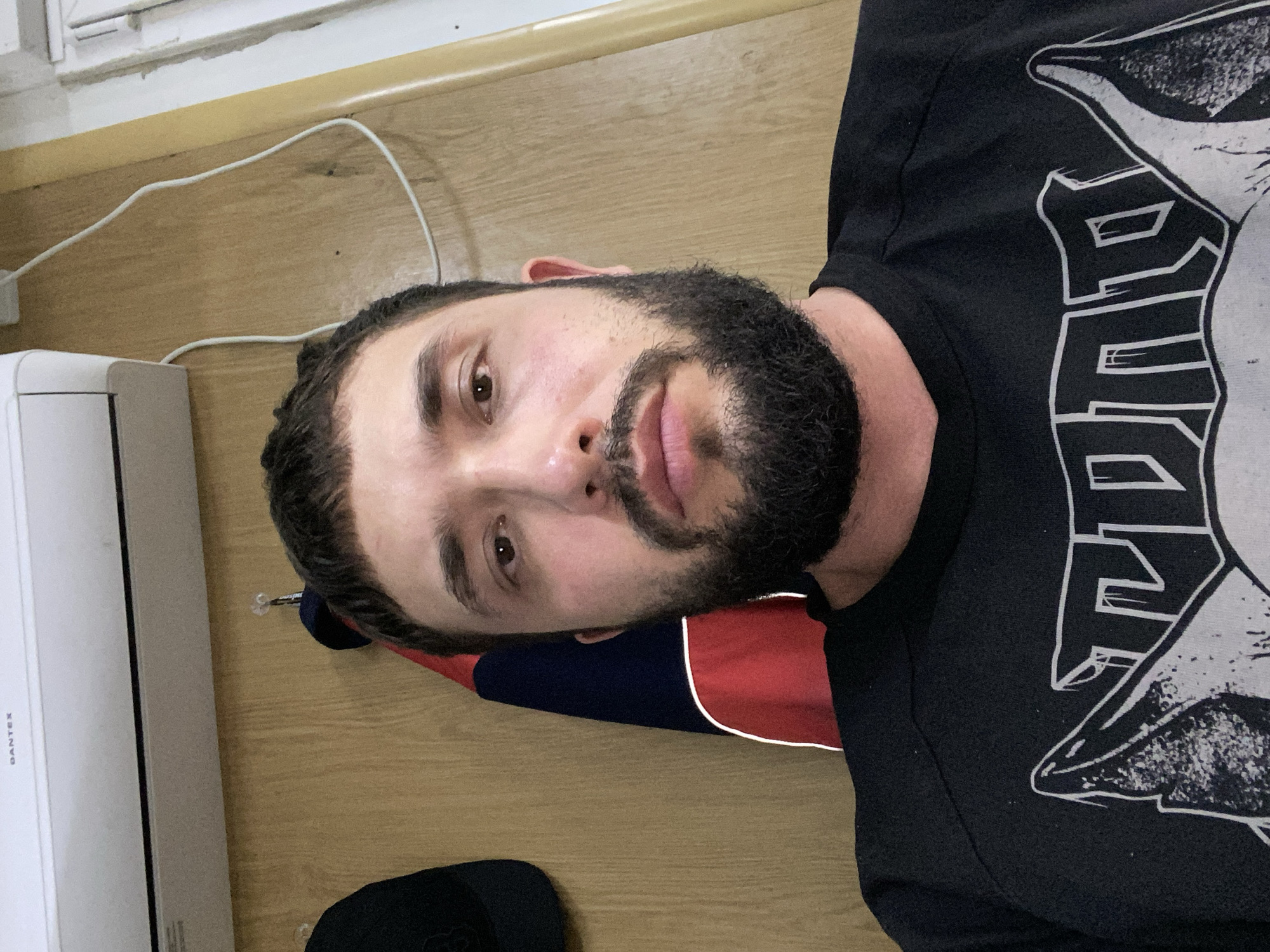 A man with dark hair and a beard is pictured indoors, facing the camera. He is wearing a black t-shirt with a graphic design. Behind him, a wooden wall is visible, with an air conditioning unit mounted high on the wall. A dark-colored cap is hanging on the wall to the left of the man, and a red and blue garment is hanging to his right. A white cord runs along the wall. The scene appears to be a domestic or personal space. The image does not contain any specific landmarks or environmental cues to determine its location within Luzhitsy, Russia.