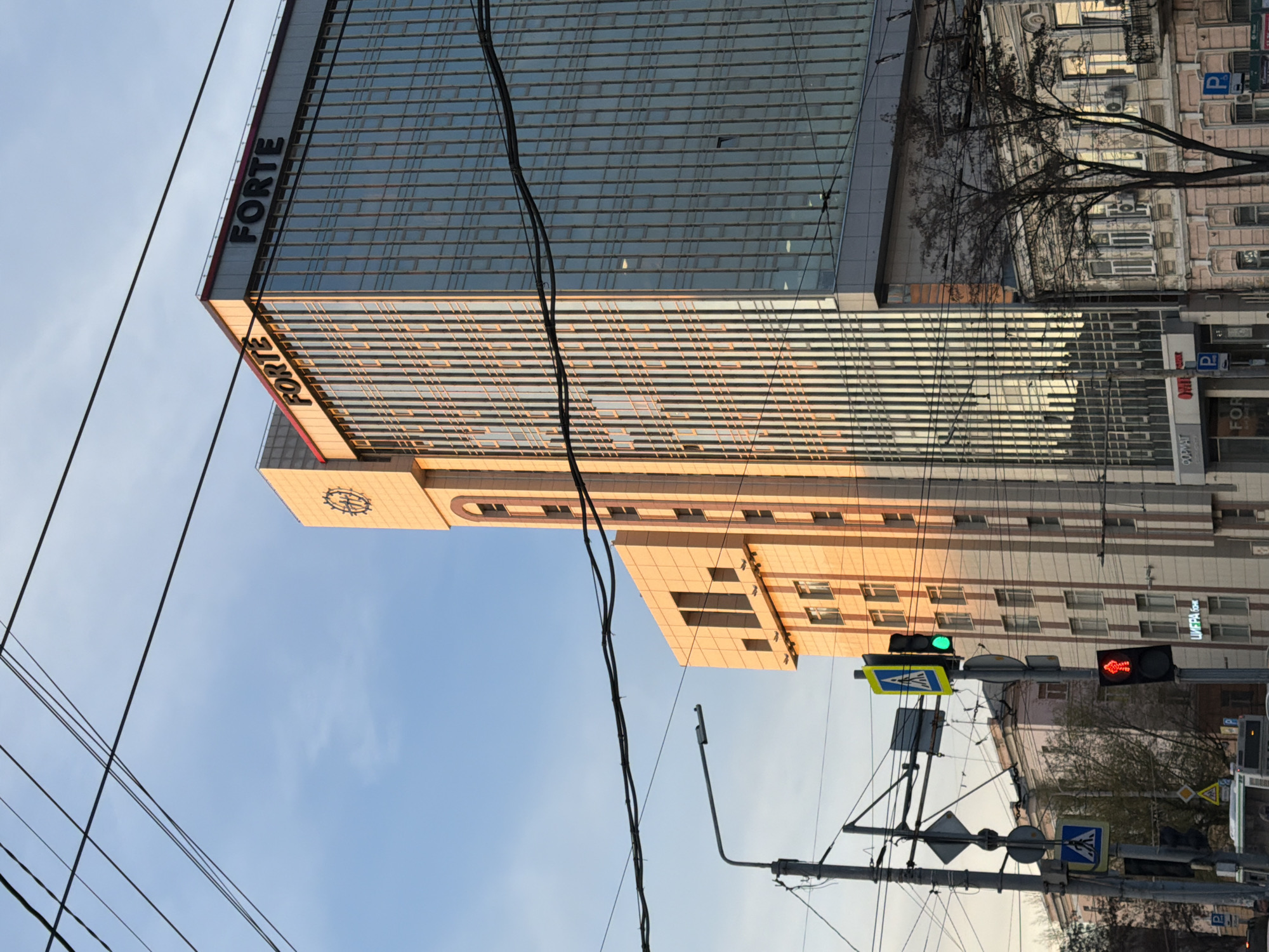 A street scene in Rostov-on-Don, Russia, featuring the "Forte" building prominently in the background. The building consists of two main sections: a lower, tan-colored block with a clock face near the top, and a taller, modern glass-and-steel structure above it, bearing the "FORTE" sign. Power lines cut across the view. A pedestrian crossing is visible with a green traffic light illuminated. Other traffic signs are visible on a metal pole. A smaller building with windows and what appears to be parking signs is seen further behind the Forte building. A few bare trees are also visible in the shot. The sky is light blue.