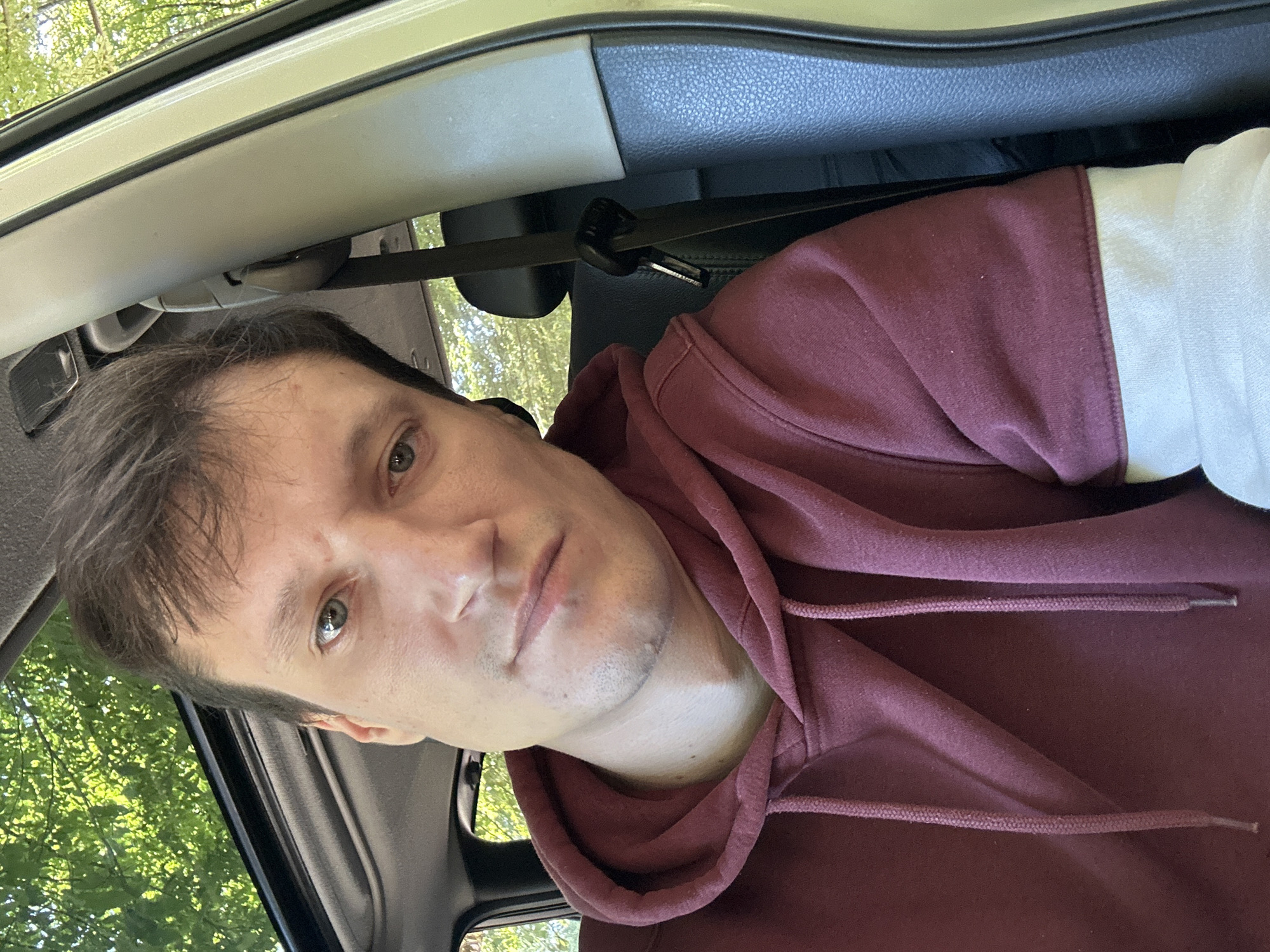 A man is captured in a close-up, selfie-style photograph taken inside a vehicle. He is seated in the driver's seat, with the sunroof open, allowing sunlight to filter through lush green trees above. The man is wearing a maroon hoodie with white accents on the sleeves. His gaze is directed towards the camera with a neutral expression. The interior of the car is visible, including the gray dashboard, a black seatbelt, and the textured gray interior paneling. The scene appears to be outdoors, as indicated by the presence of trees and natural light. The location is identified as Shatava, Ukraine.