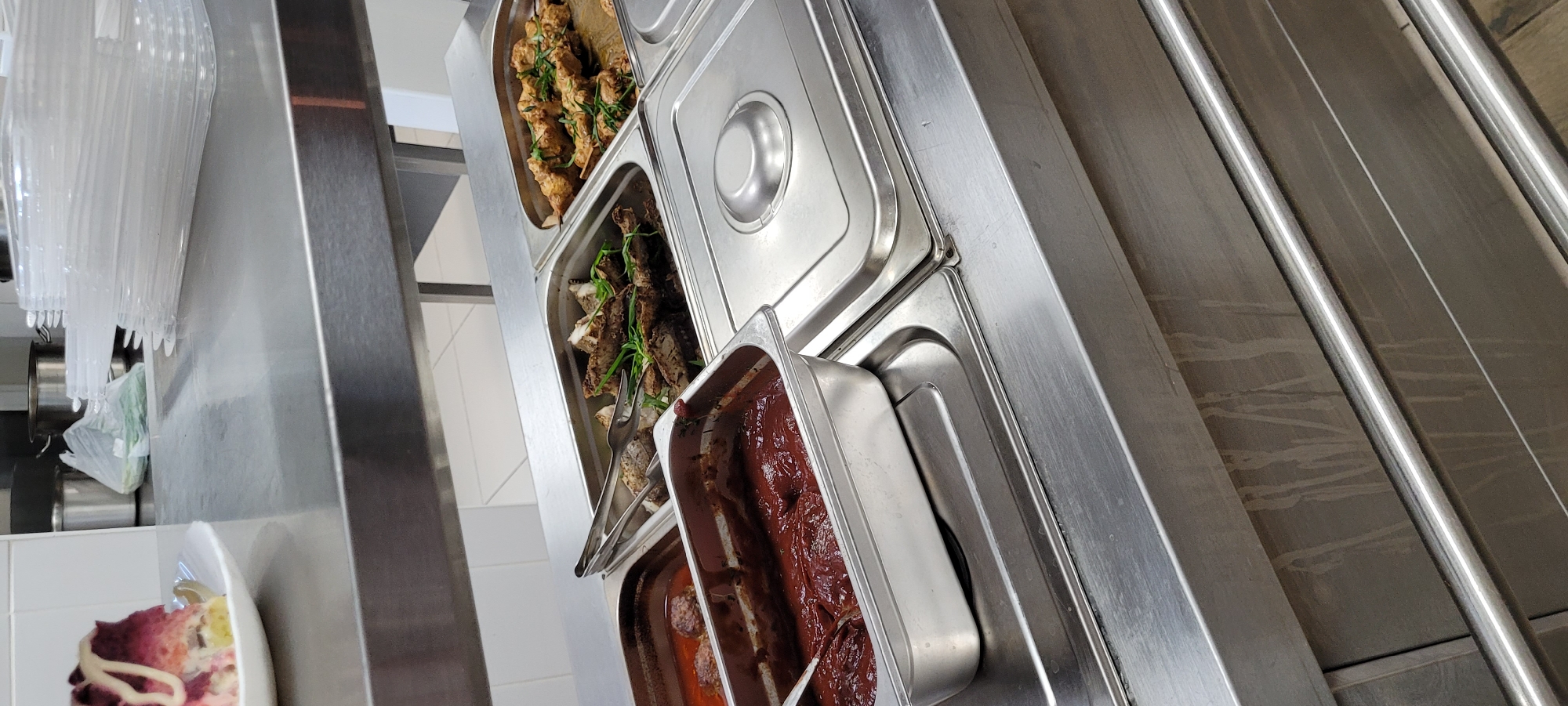 The image shows a metal serving counter with four trays of food. The topmost tray appears to contain skewers of cooked food, possibly chicken or meat, garnished with green herbs. Below that is a tray of cooked fish with green herbs and a pair of metal tongs resting on top. The next tray has a dark red, possibly meat-based, sauce. The bottom tray is partially visible and seems to contain food items covered in a similar sauce.
To the left of the counter, there's a stack of clear plastic serving utensils and a plate of food with a white sauce on top. The surroundings are primarily stainless steel, suggesting a commercial kitchen or cafeteria setting. The location is Unknown, Unknown.