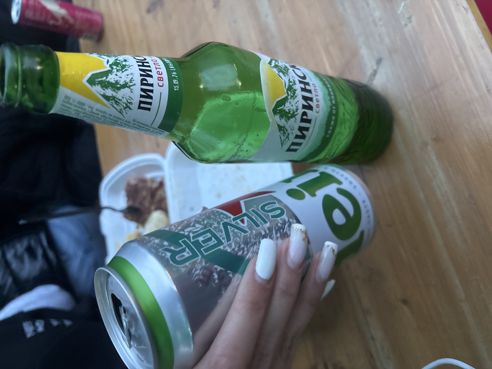 The image displays a casual setting featuring alcoholic beverages and a partially visible food container on a wooden surface, likely in Plovdiv, Bulgaria. In the foreground, a person's hand with long white, square-tipped nails, accented with gold glitter on one finger, holds a silver can. The can has the word "SILVER" prominently displayed on its side in white and green lettering. The top of the can is open.
Above the can, a green glass bottle is lying horizontally on the wooden surface. The bottle's label, yellow and green with a mountain peak graphic, is clearly visible. The Cyrillic text "ПИРИНС" (PIRINS) and "светло" (light) are visible, indicating the brand and type of beer. Another part of the label includes "15.09.26 LN60UP!", which likely indicates a best-before date. Additional text reads "СВЕЖО ОТ ПЛАН..." (Fresh from the plan...) and "одовж стикета на гърлото. Тел: 0000 11 500" (along the neck label. Tel: ...).
In the blurred background, another red can, possibly a soft drink or energy drink, is visible. A white styrofoam container with some food inside and a plastic fork is also present, suggesting a meal is being consumed. The wooden surface appears to be a table. The lighting suggests it is daytime.